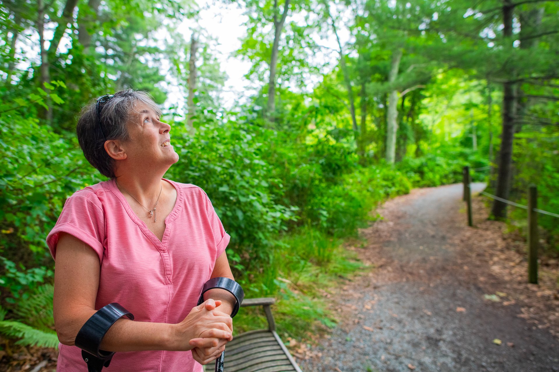 A woman in a pink shirt using forearm crutches looks up into the lush green canopy, pausing to rest at a bench along Stony Brook's wheelchair-accessible All Persons Trail