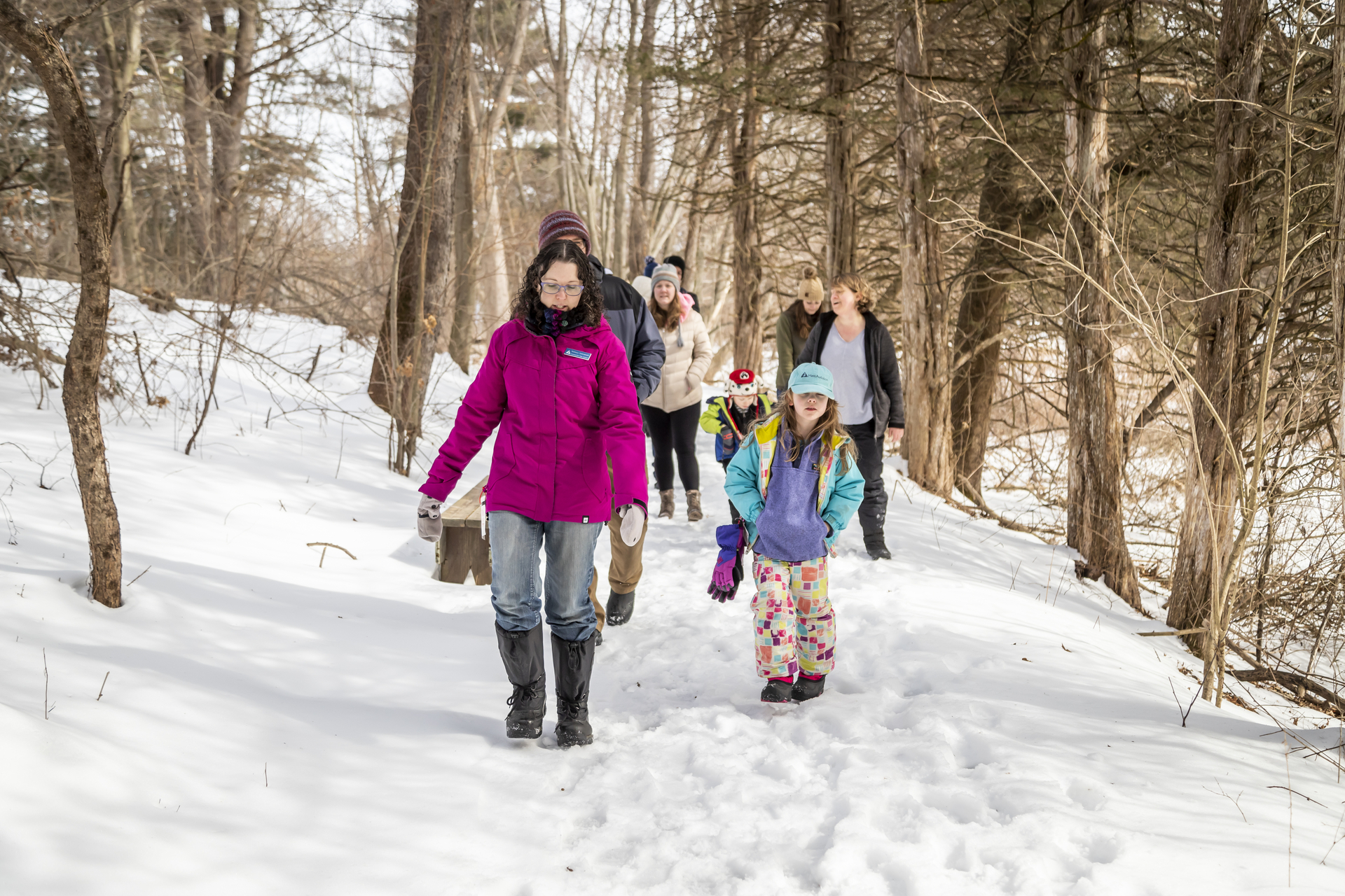 Group of people walking through the snowy woods. A woman in a pink jacket and a young girl with a teal jacket and hat.
