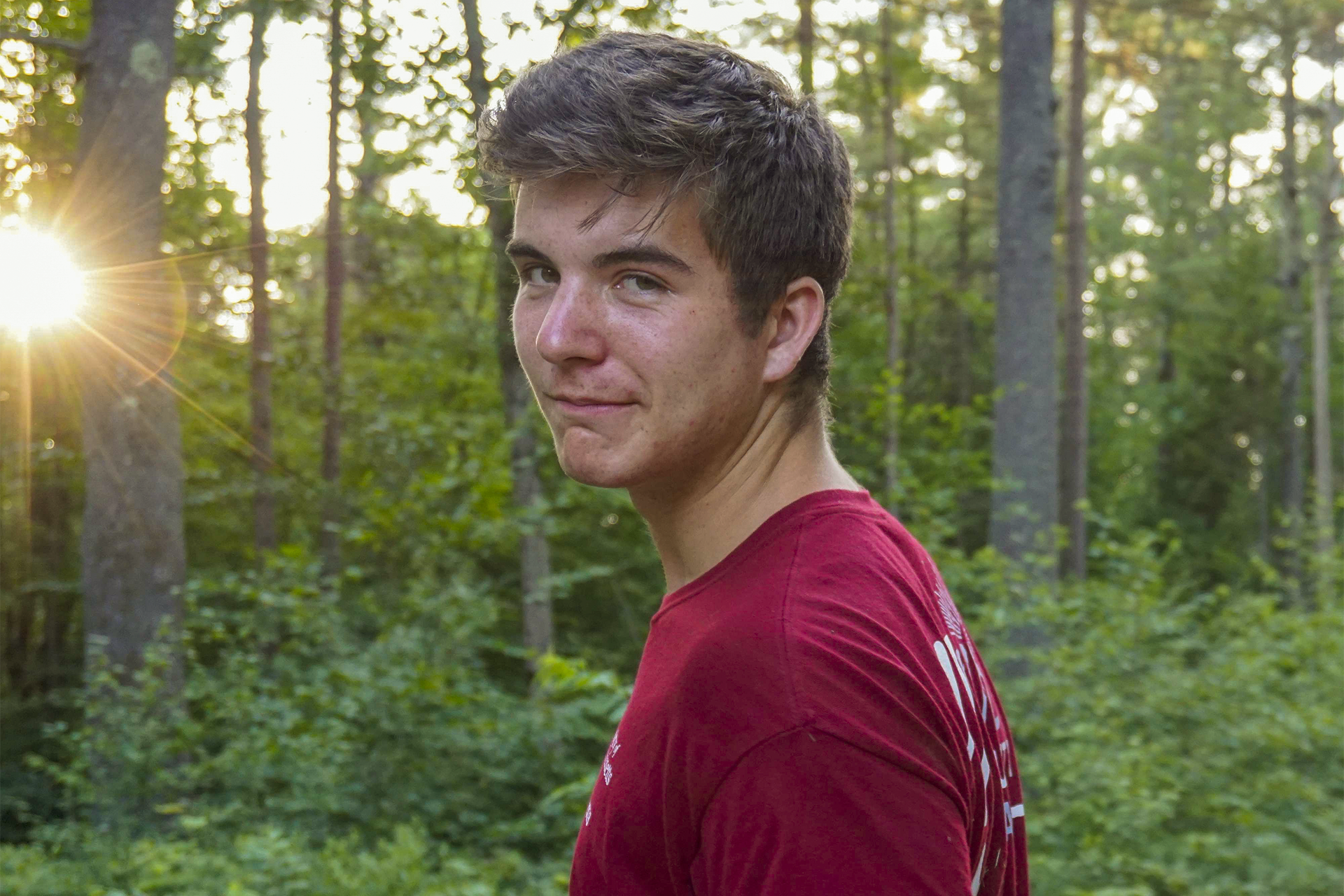 Brunette boy smiling at the camera in a red shirt. The sun filters through the trees in the background.