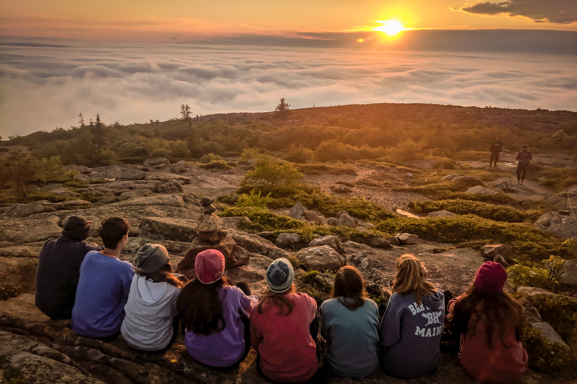 A group of eight teens sits in a row with their backs to the camera, gazing out across a rocky mountain summit with few trees. In the distance, the sun sets over a low-lying bank of fog, which looks like a sea of clouds.