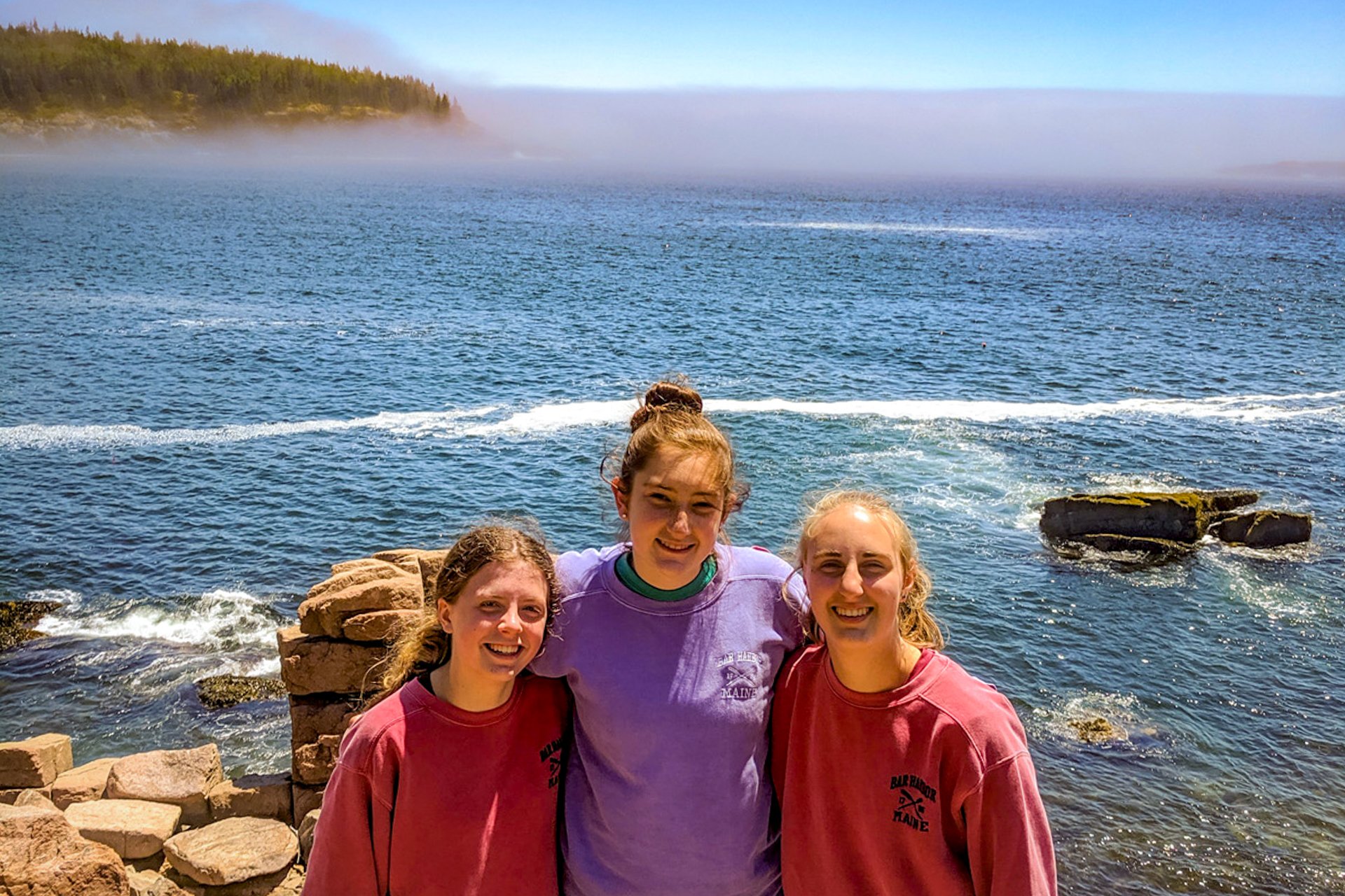 Three teen girls pose in front of a rocky shoreline overlooking the ocean, somewhere along Maine's rugged coast