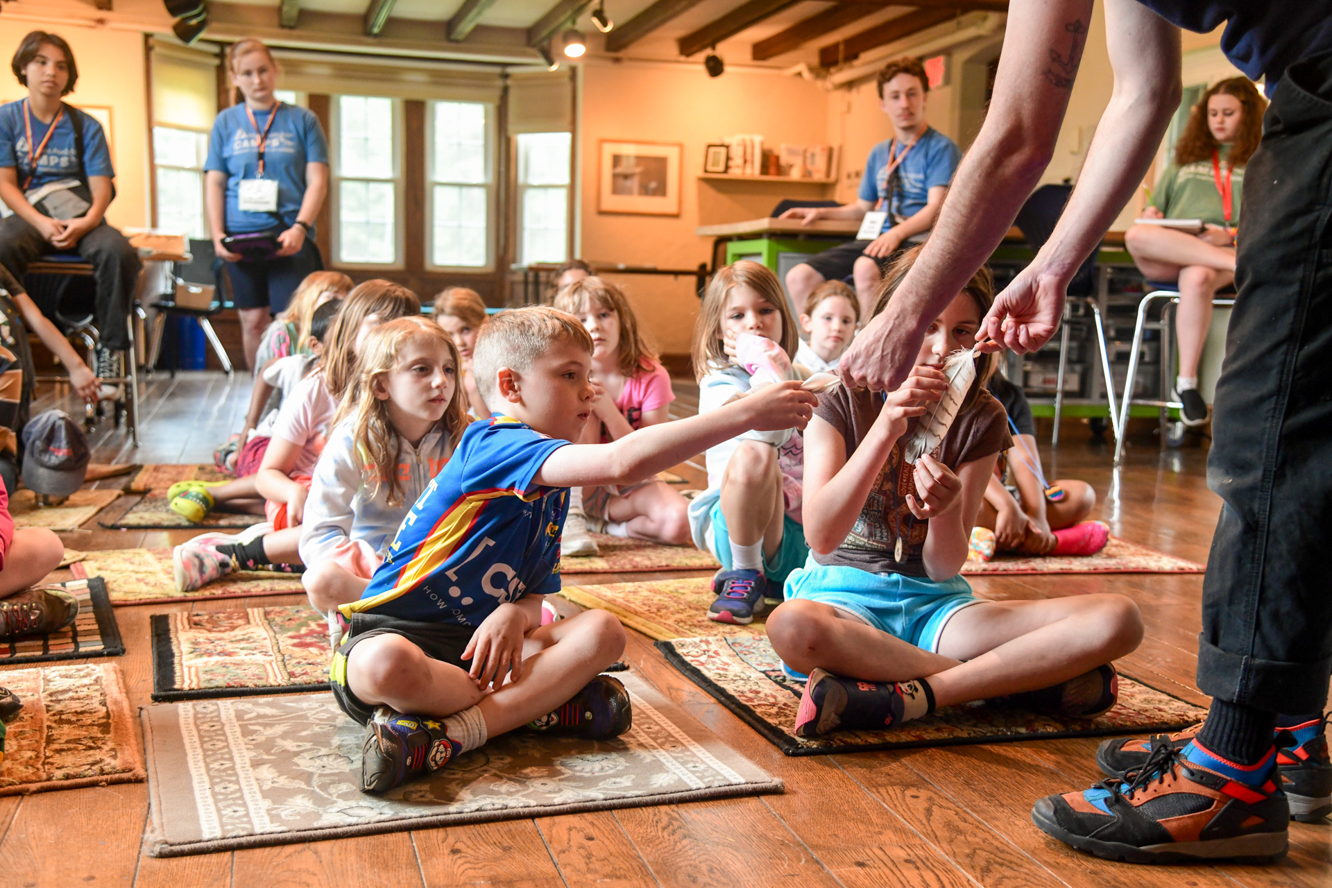 MABA campers seated cross-legged on small indoor rugs, reaching for bird feathers being held out by a teacher naturalist