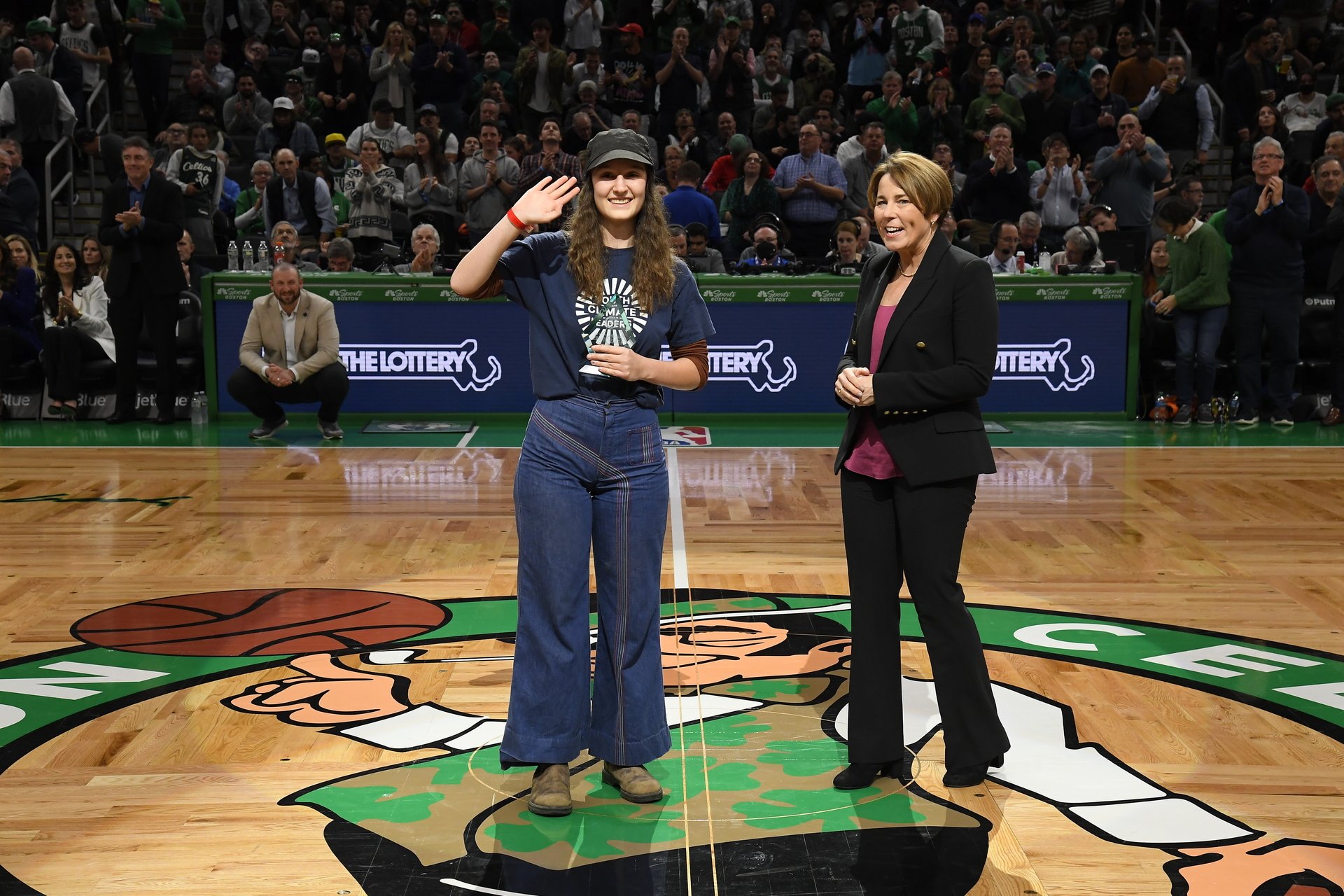 Young girl in jeans waving at camera while another woman stand next to her in a pant suit.