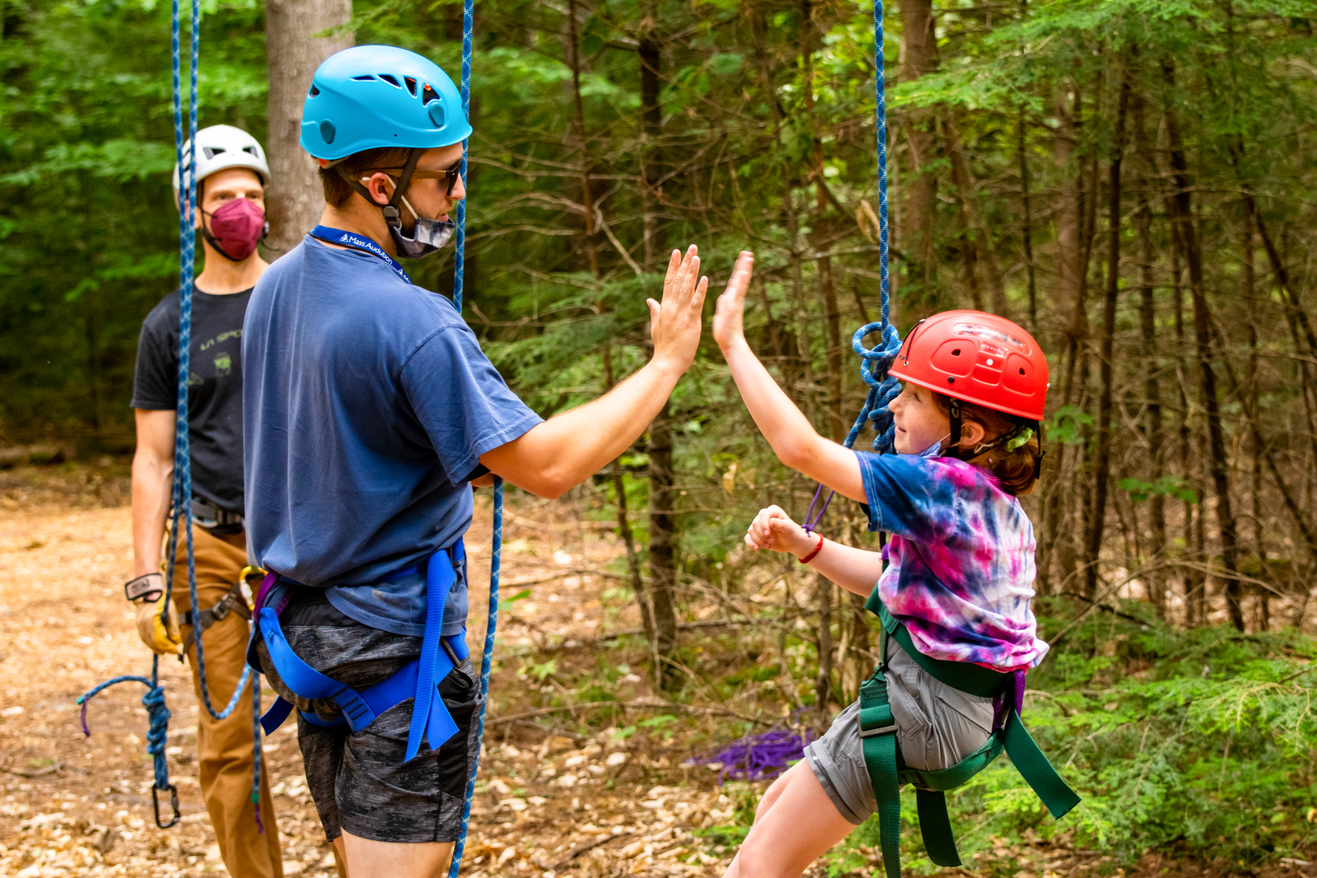 A Wildwood camper and counselor exchange a high five at the high ropes course. Both are wearing climbing helmets and harnesses, and the camper is hanging just above the ground.