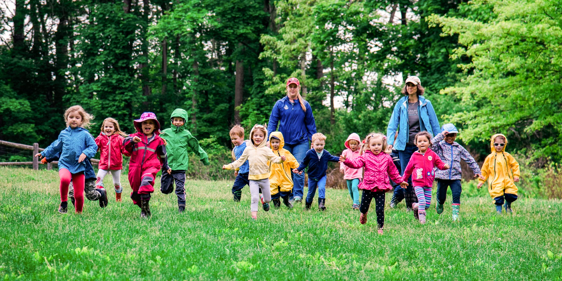 A group of preschoolers at Drumlin Farm Community Preschool running across a field surrounded by forest, with two teachers smiling and walking behind them