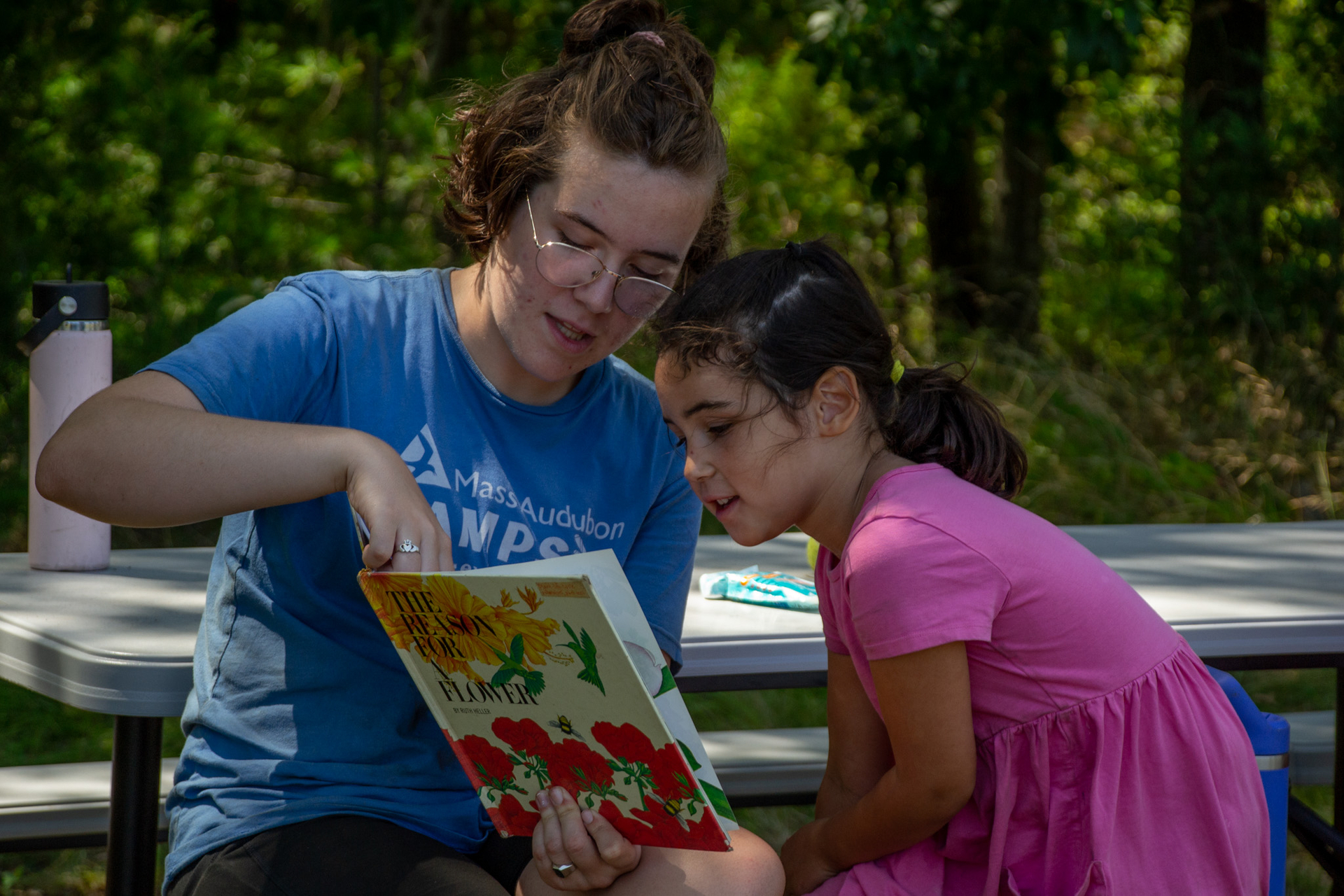 A counselor points at a picture book while a small camper sits next to her and looks on.