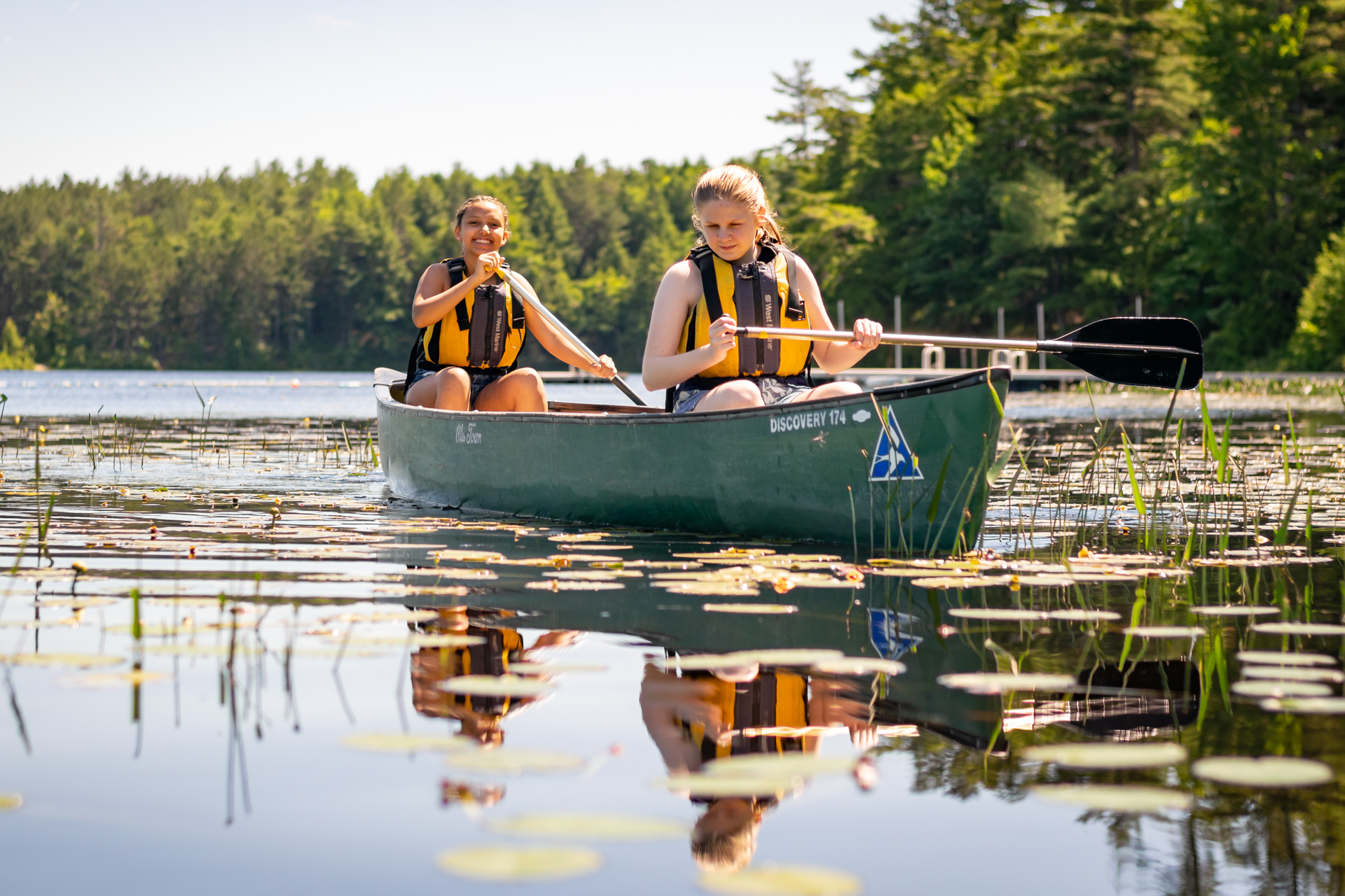 Two campers wearing yellow life jackets and paddling through lily pads in a green canoe with a Mass Audubon logo on the prow, their smiling faces reflected on the still water below