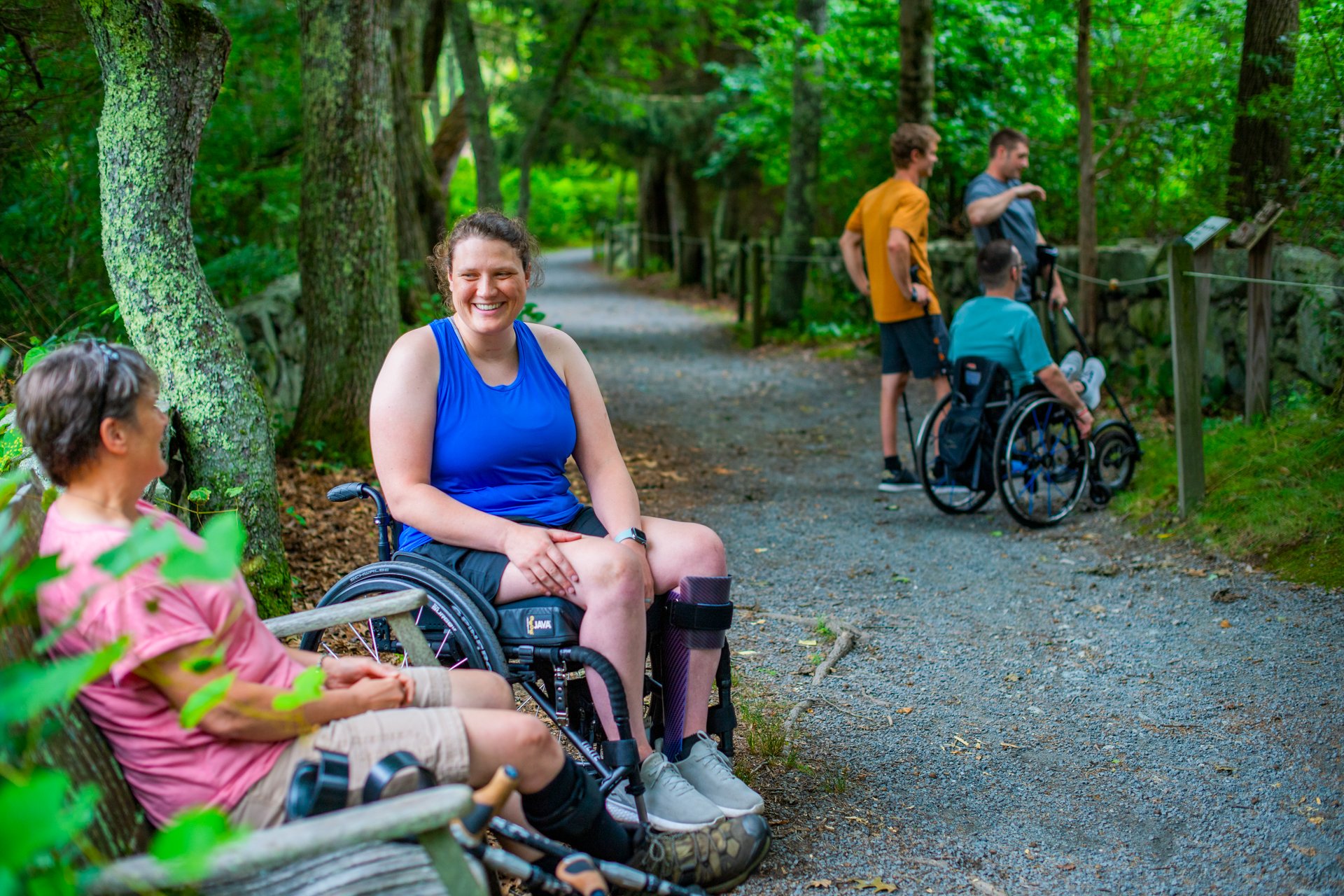 A group of people with disabilities, using wheelchairs, forearm crutches, and other mobility devices enjoying the wheelchair accessible trail at Stony Brook Wildlife Sanctuary in Norfolk