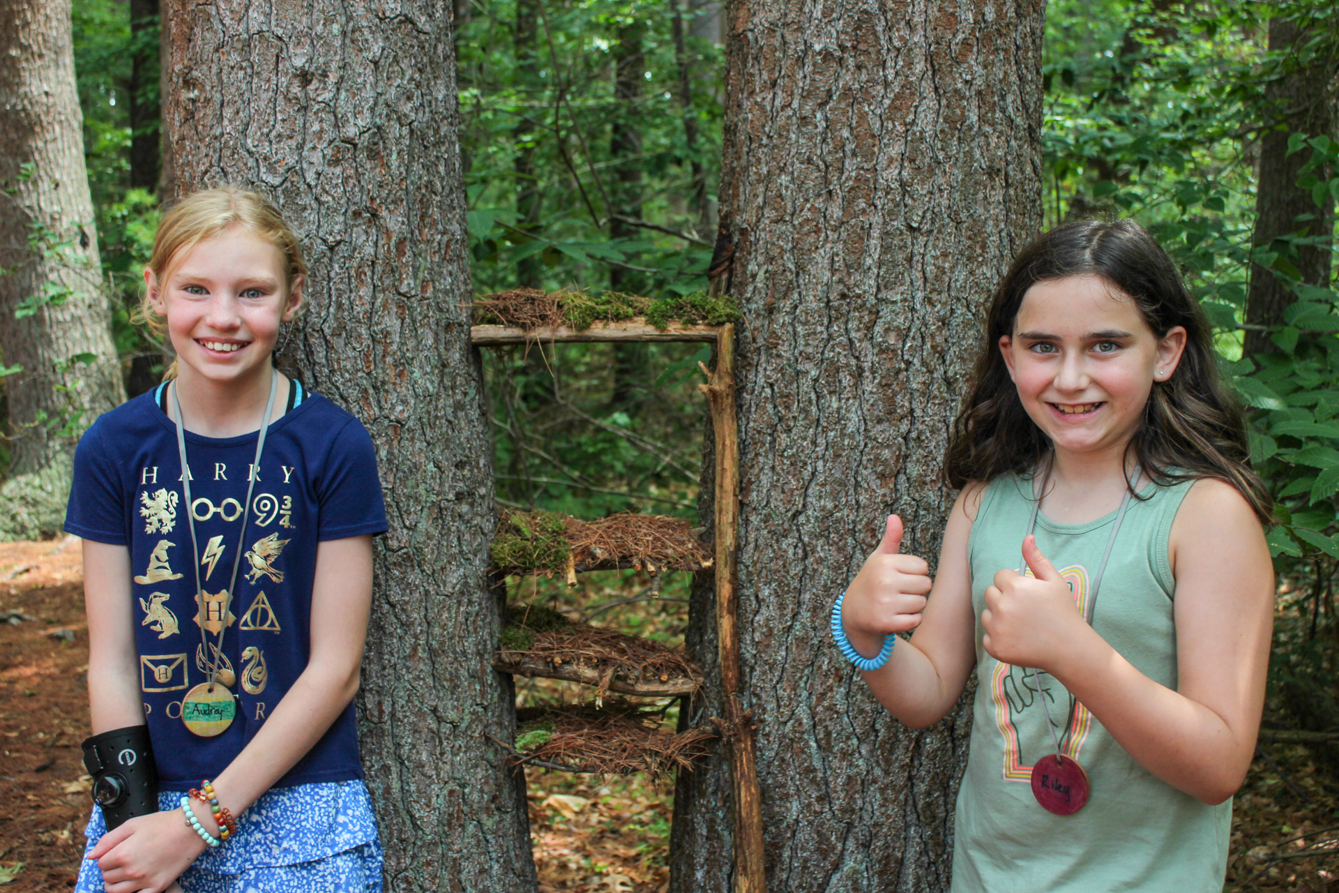 Two Broadmoor campers standing in front of two pine trees, between which they have built a multi-tiered structure of sticks, moss, and pine needles. Both children are smiling and one has two thumbs up.