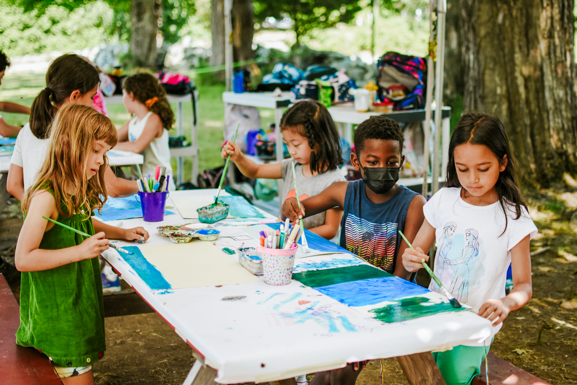 Campers at MABA's Wild At Art Nature Camp painting at a table beneath the shade of a large tree