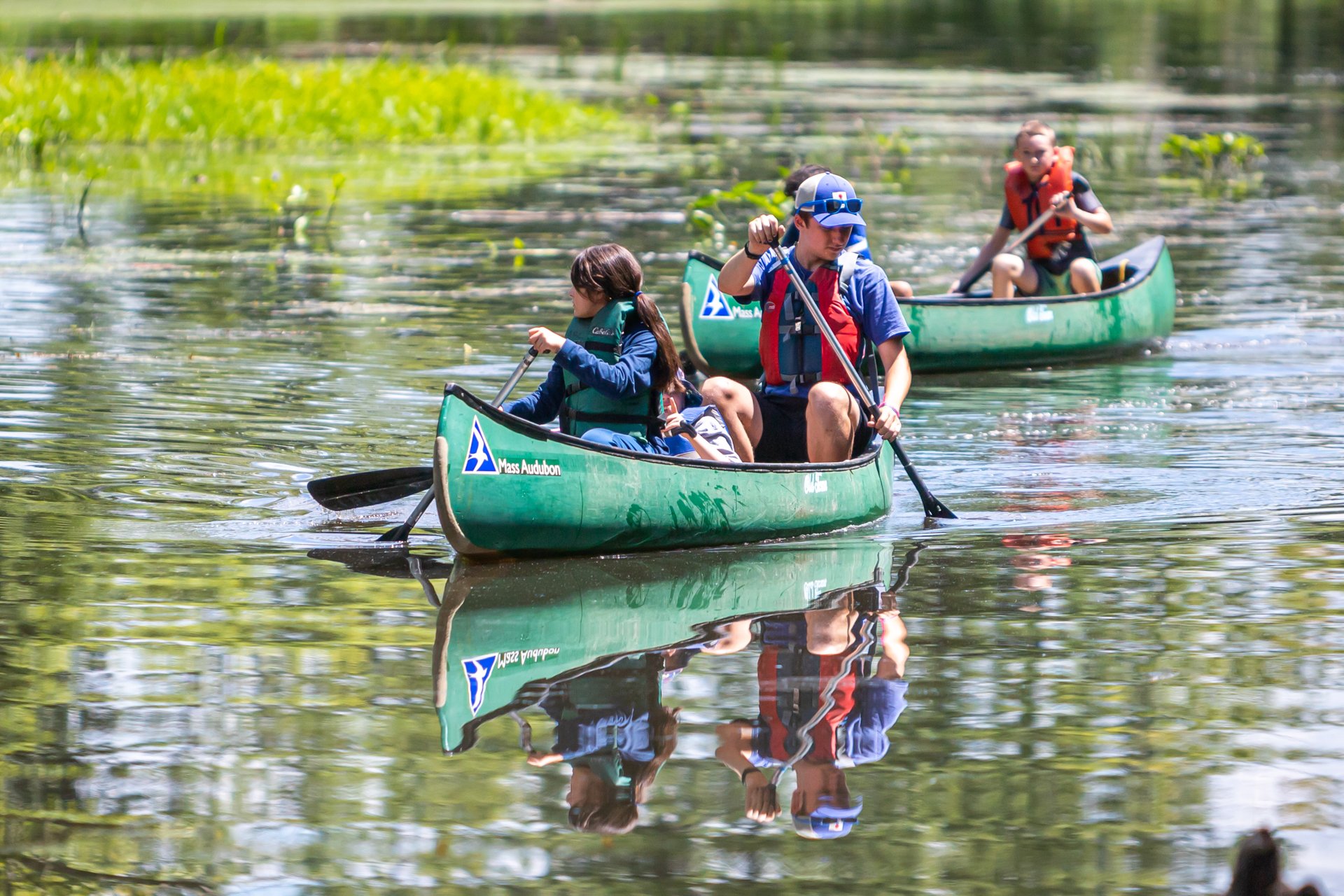 Campers and a counselor at Arcadia Nature Camp paddling canoes marked with the Mass Audubon logo down a peaceful river