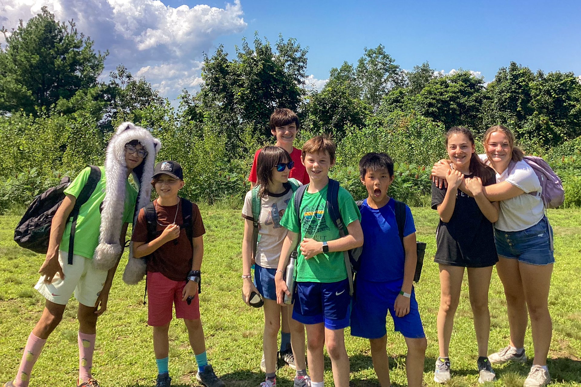 A group of Blue Hills campers poses for the camera with smiles; one is wearing a silly faux fur hat and a few have their arms around each other in side hugs.