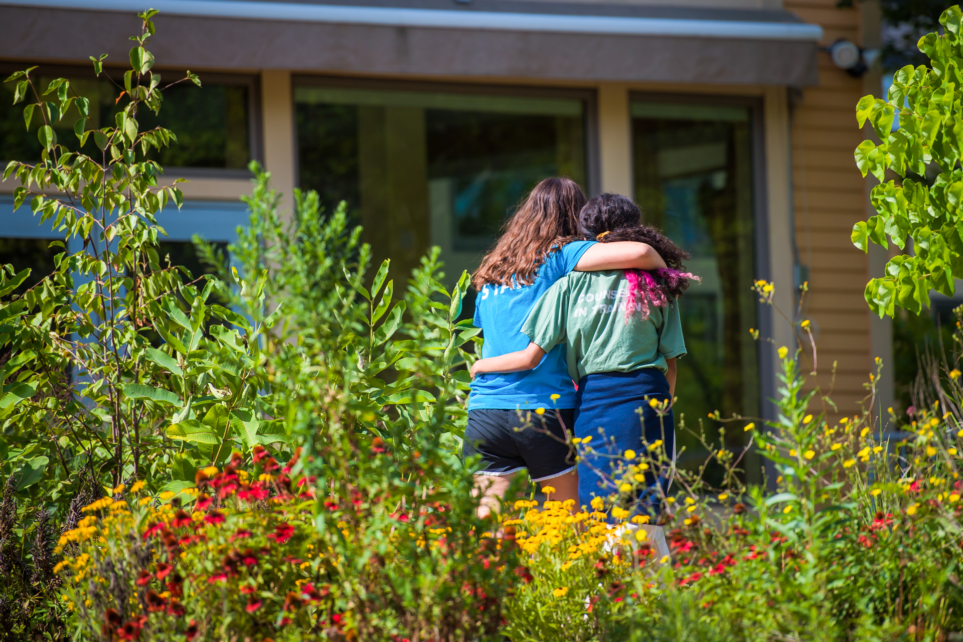 A counselor and CIT with arms around each other walking through the sanctuary's pollinator garden, filled with native wildflowers