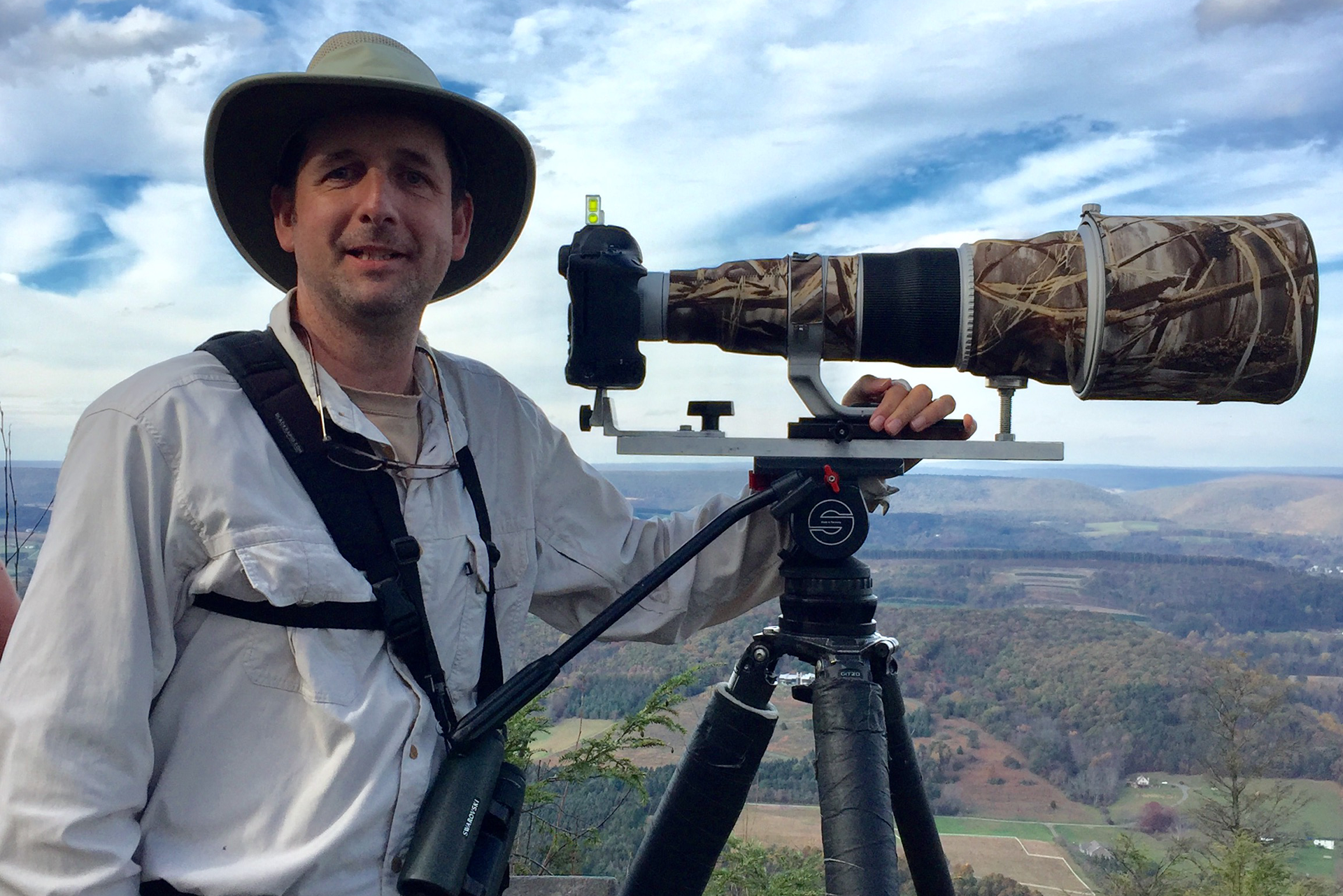 Man with a hat stands smiling next to a large, camouflaged scope.