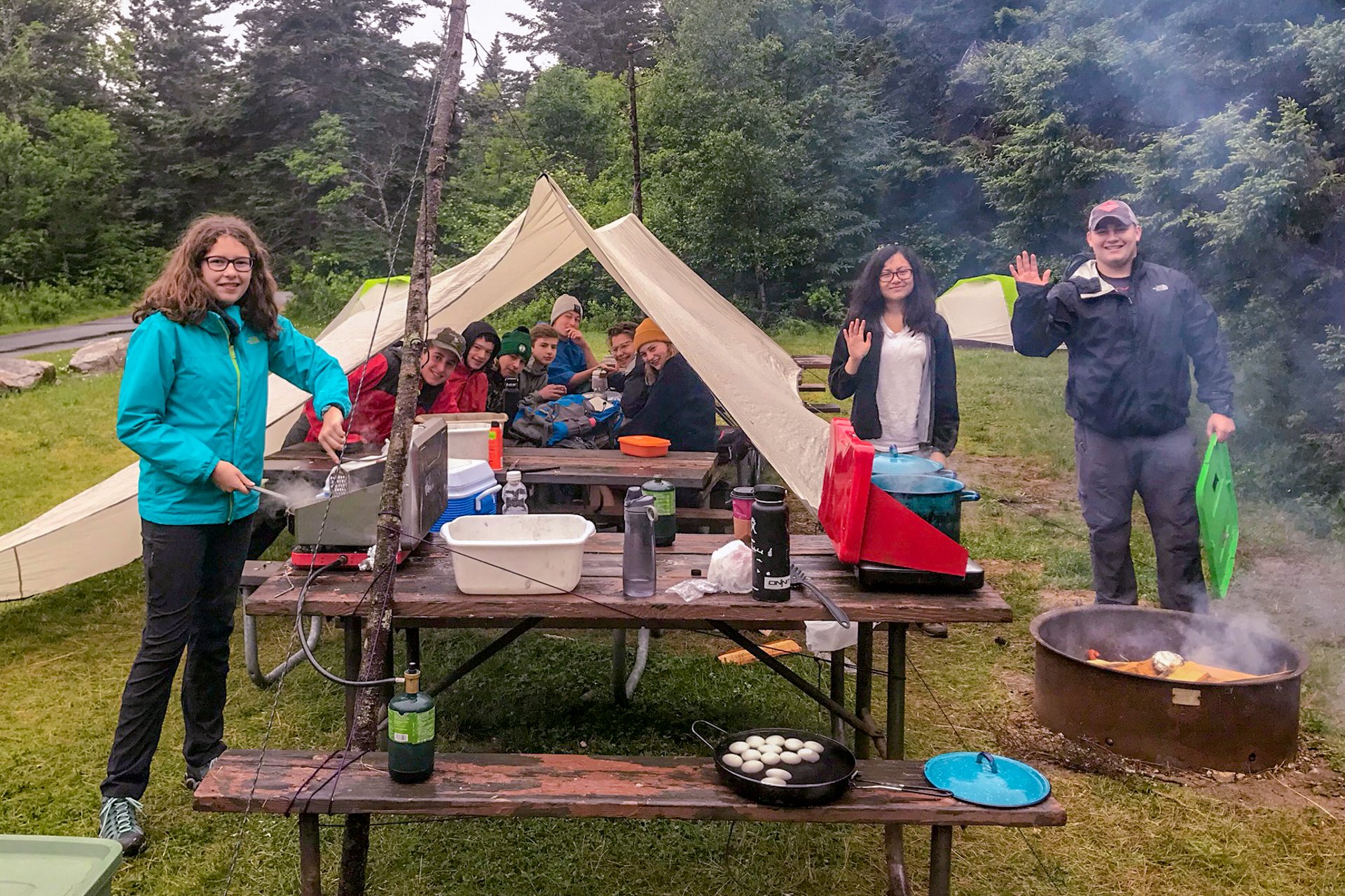 Wildwood teen adventure trip campers (trekkers) at a campsite with a tent shelter, picnic table, and a fire ring, cooking a meal together
