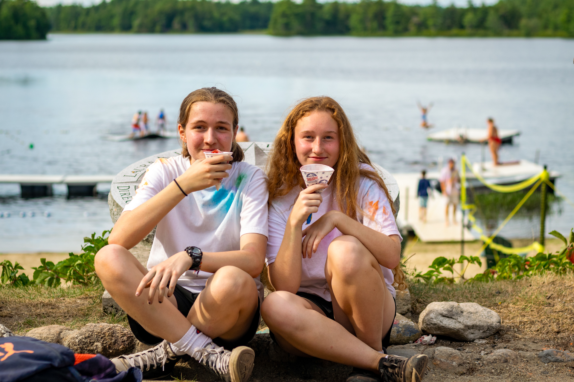 Two girls, about 12 years old, are seated cross-legged, smiling and holding up Sno Cones. In the background, you can see Hubbard Pond and various children swimming and jumping off the docks.