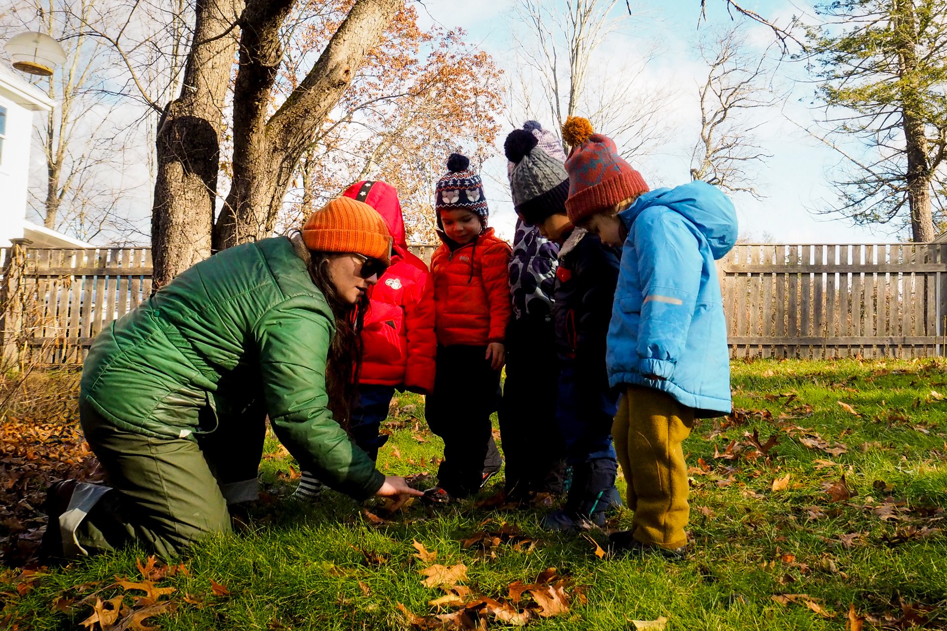 A group of preschoolers in winter coats and hats huddle around their teacher who is kneeling in the grass and pointing to autumn leaves on the ground