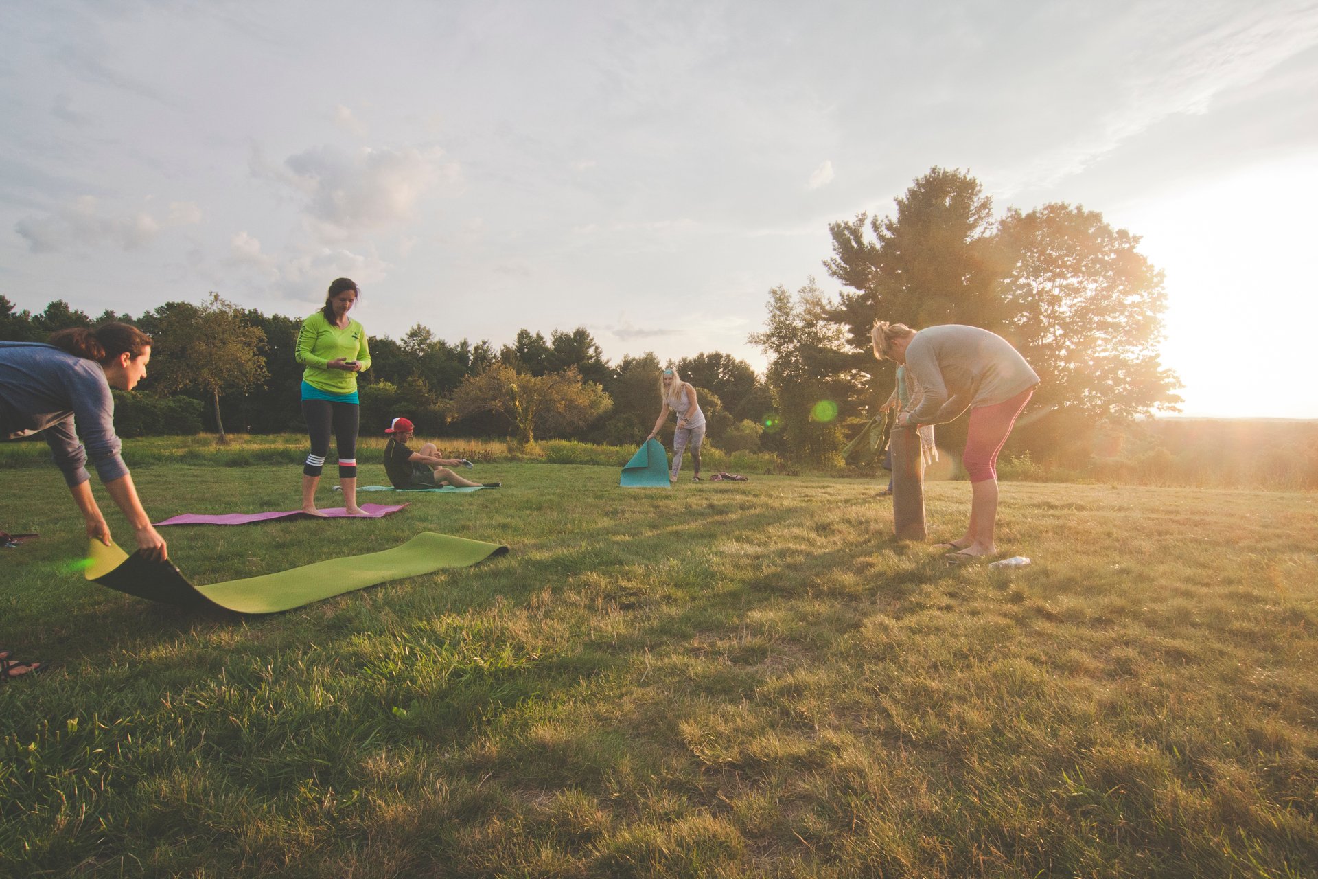 A group of five people rolling out different colored yoga mats in a flat, grassy area.