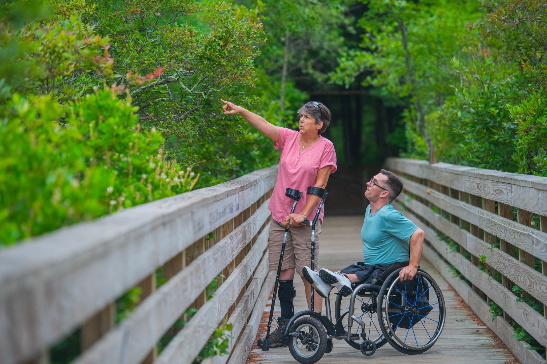 Two people on Stony Brook's accessible All Persons Trail boardwalk: One is standing, using bilateral crutches and pointing toward the tree canopy while the other, using a wheelchair, looks in the same direction.