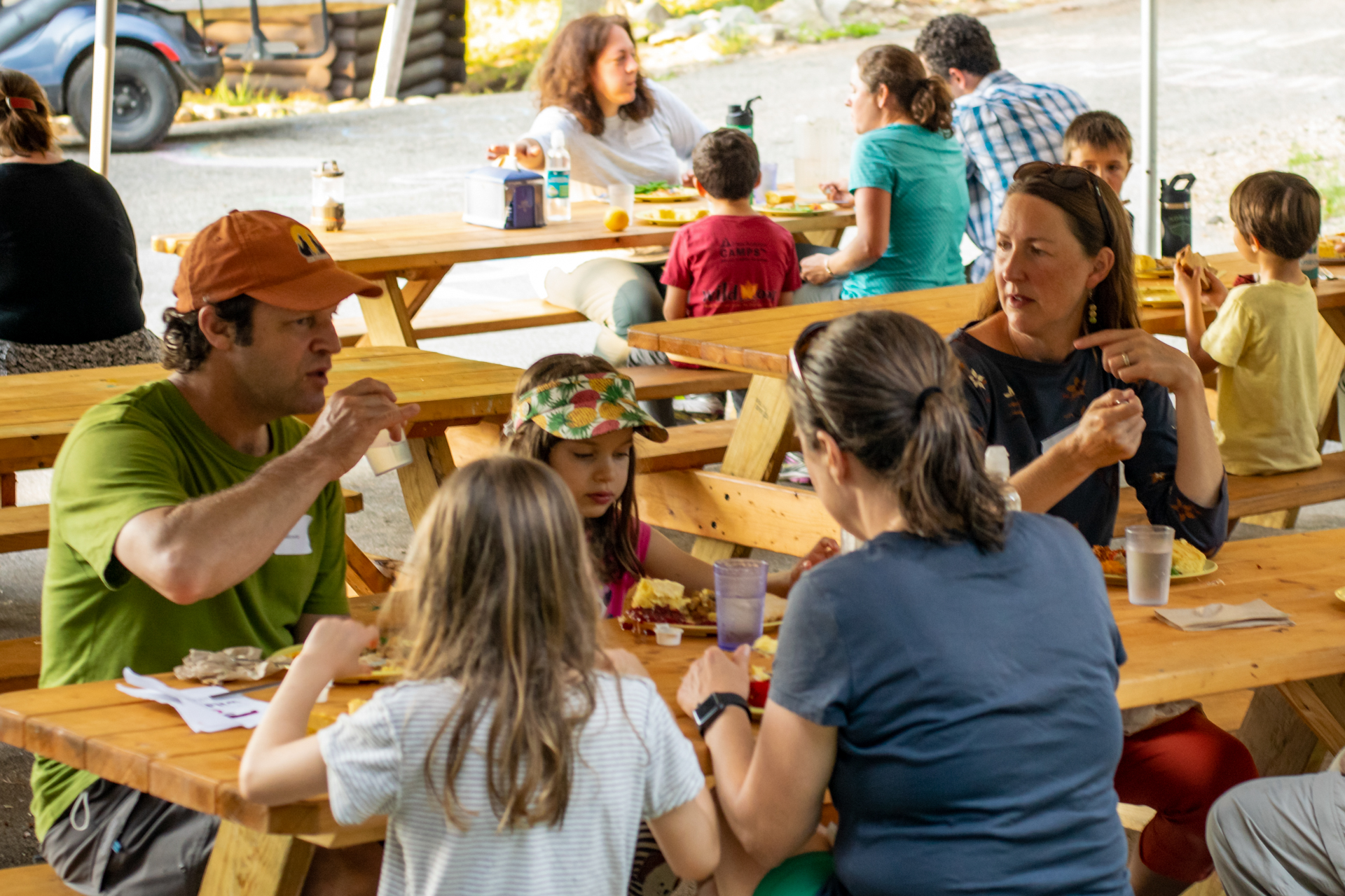 Several adults and children are seated at a group of picnic tables under a large tent, enjoying a family-style camp meal