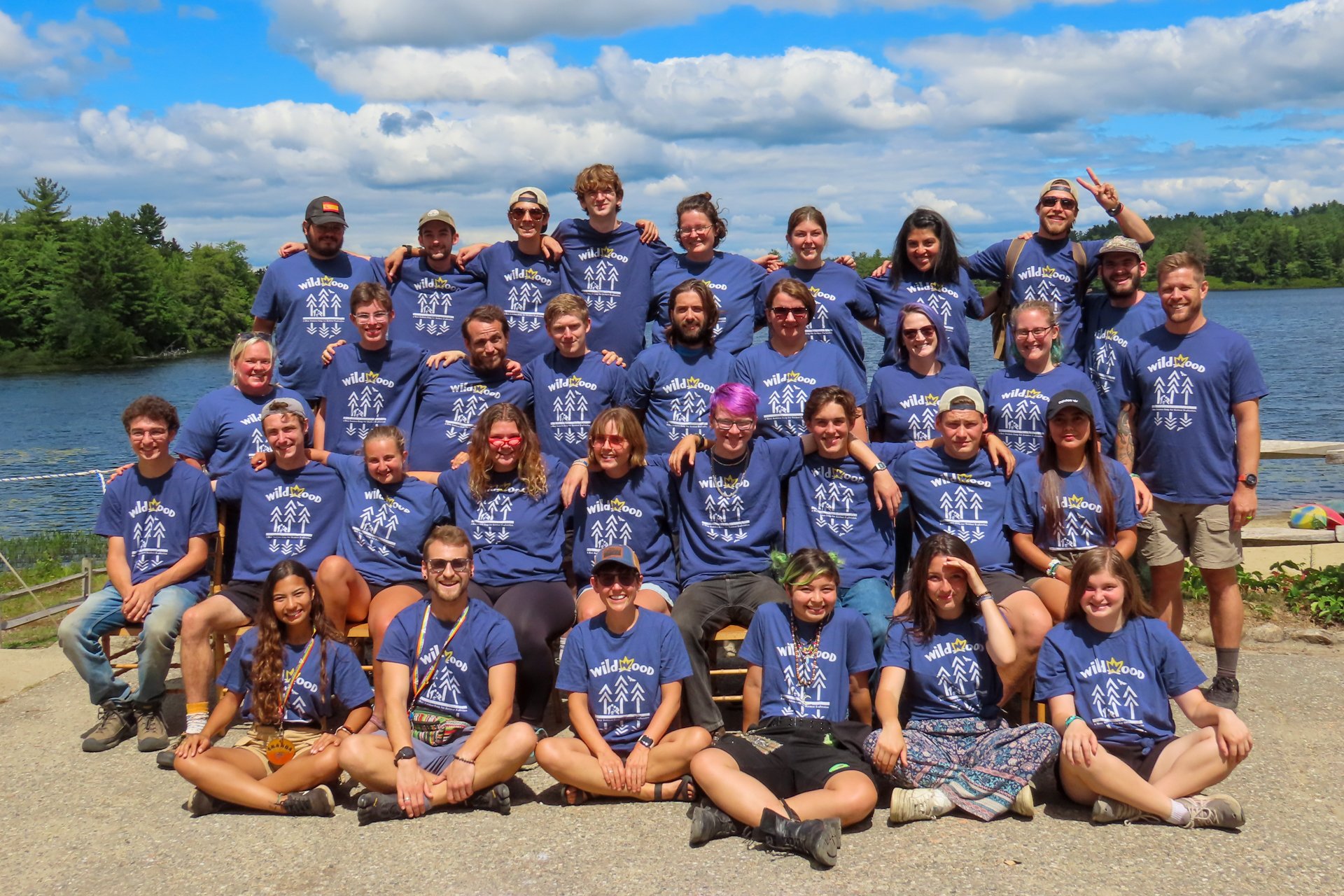 A group of Wildwood staffers wearing matching camp shirts, seated or standing in four rows in front of the camp's waterfront