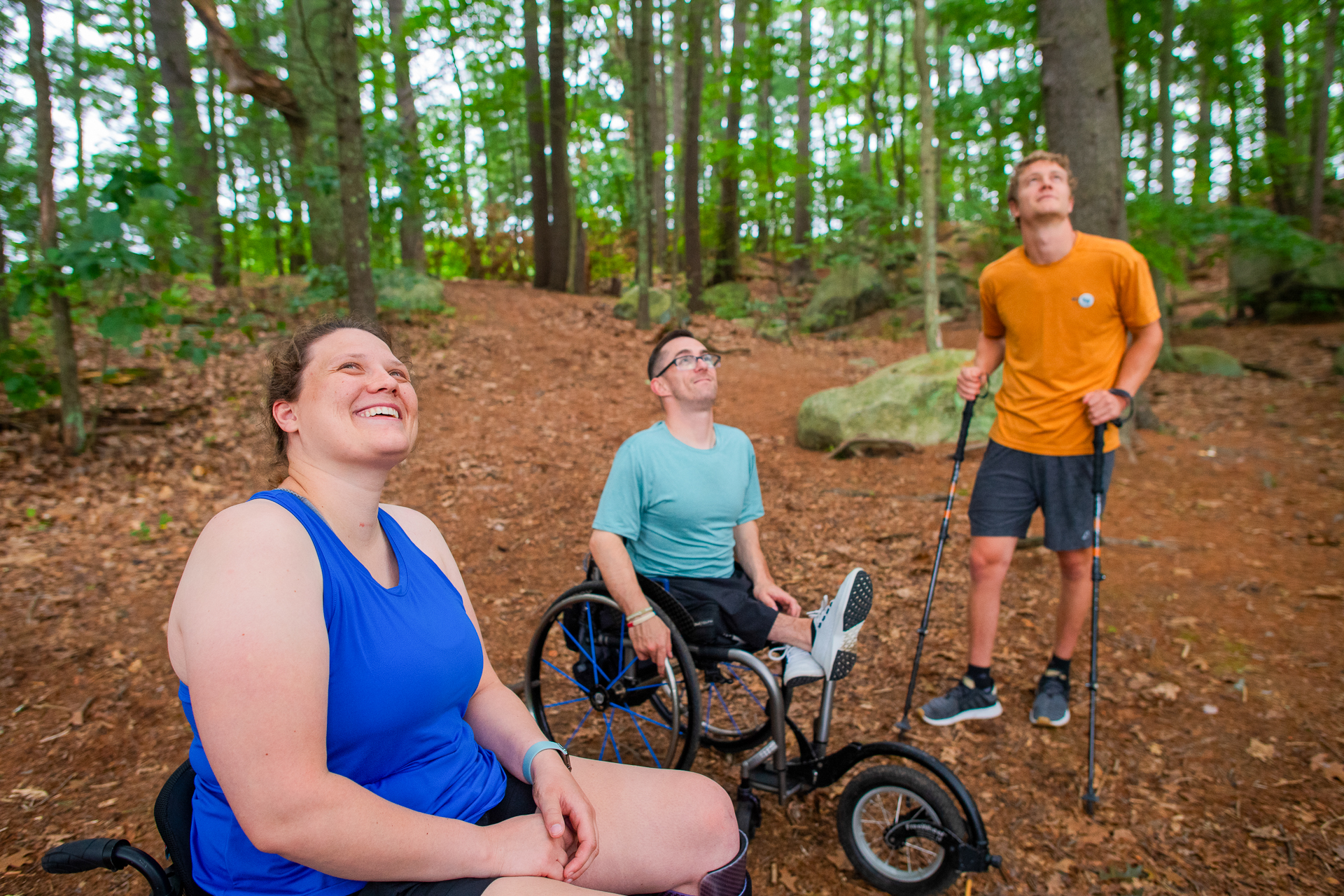 Three people in a forest, two using wheelchairs and one using trekking poles, looking up into the canopy and smiling