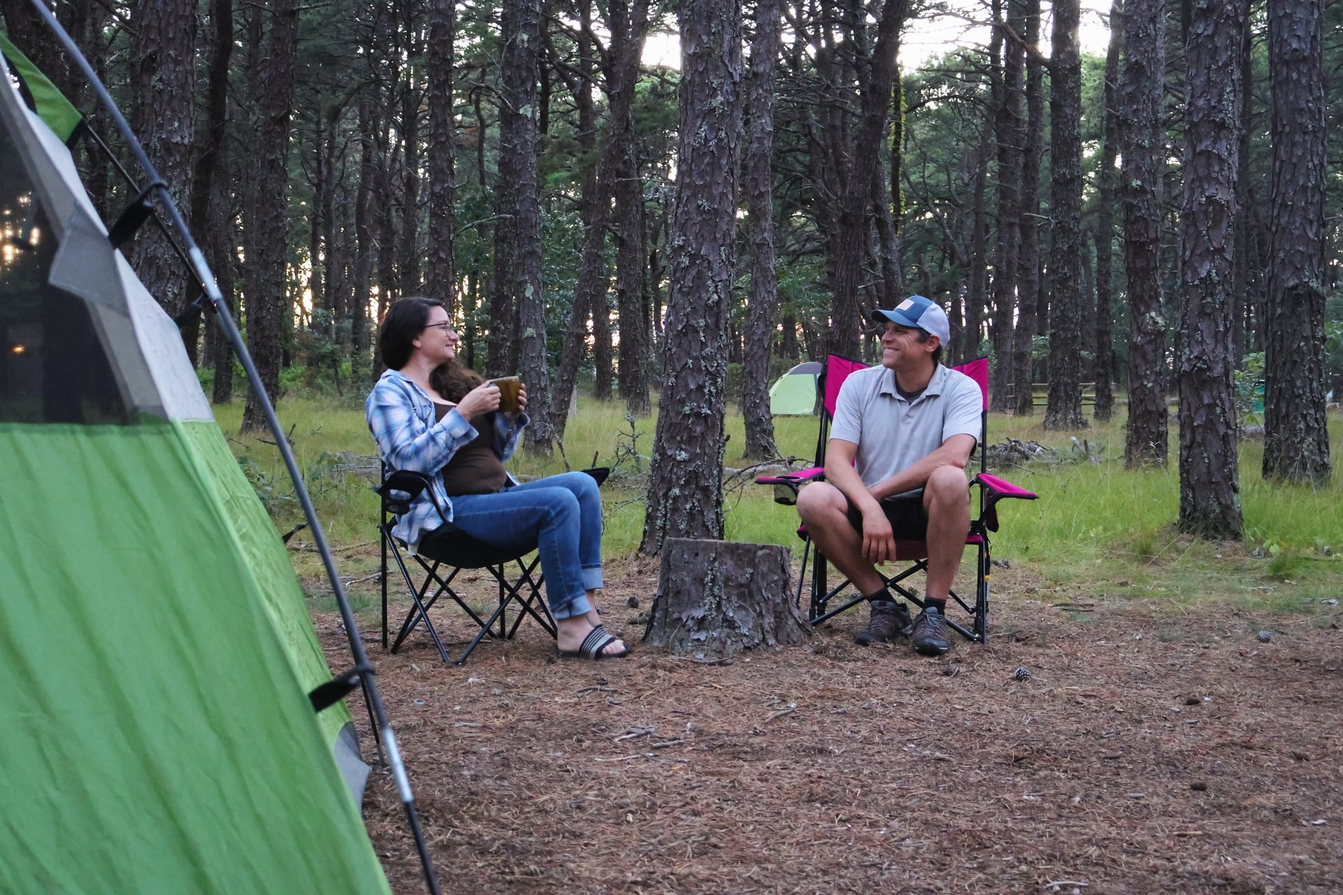 Two people sit in camp chairs at the Wellfleet Bay campground. A  tent is in the foreground, and many trees are behind them.