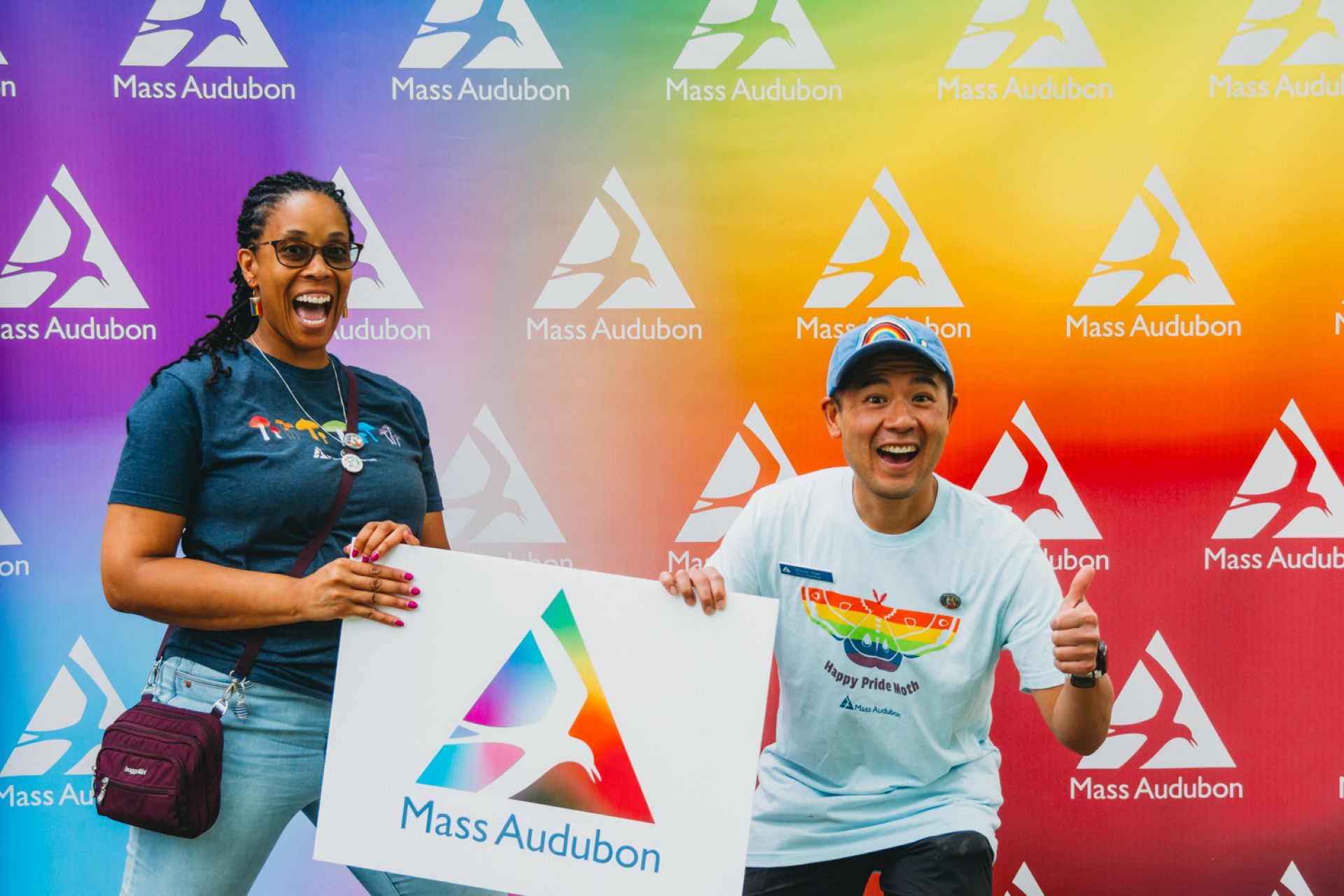 Two people smile and thumbs up with a Mass Audubon pride sign on a rainbow background.