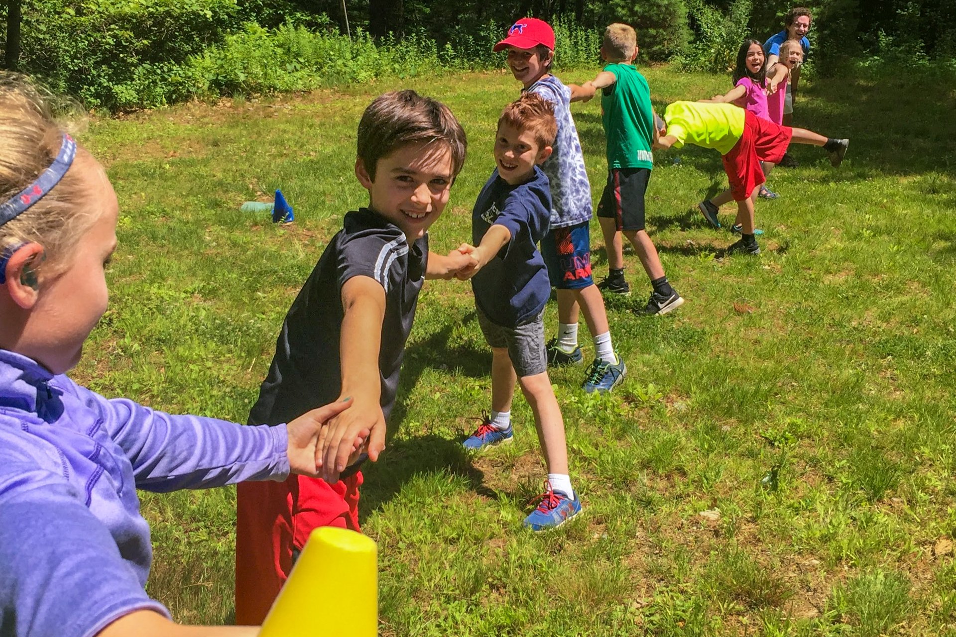 Campers at Broadmoor Nature Camp playing a game in the grass, smiling and holding hands in a line that stretches away from the camera