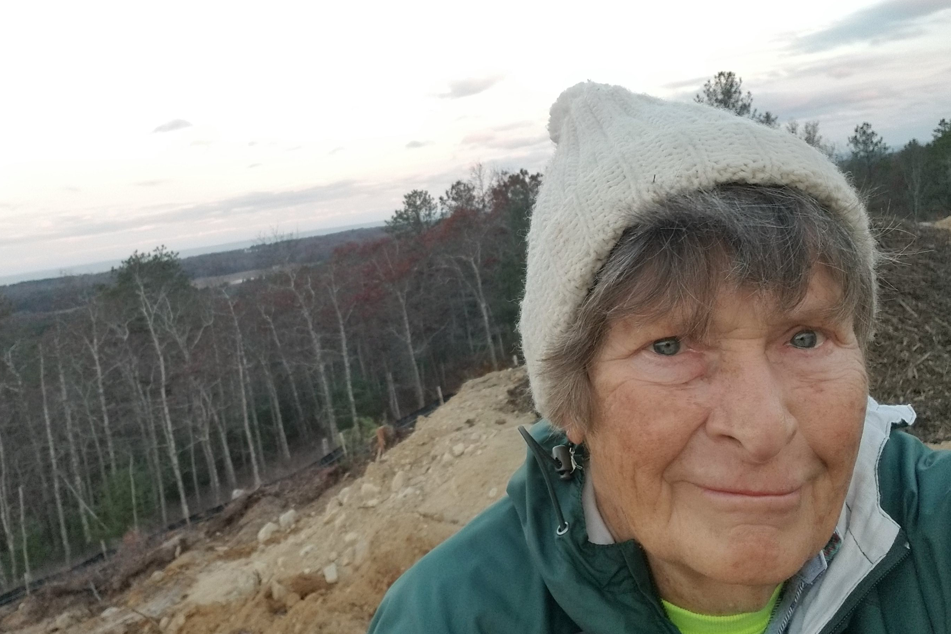 Person in a white knit hat smiles in front of a hillside with work being done on it.