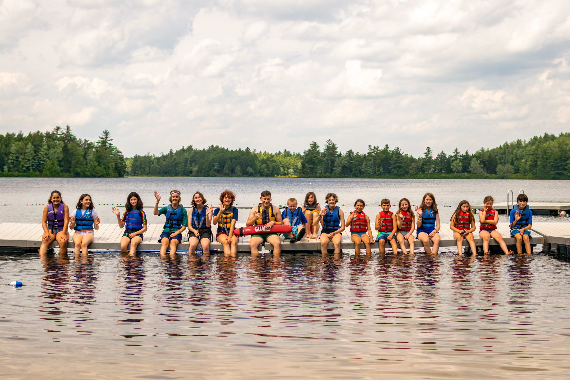 Wildwood campers wearing swimsuits and life jackets seated along the edge of the dock with their feet in the water, smiling and waving; a lifeguard holds a flotation device in the center of the group.