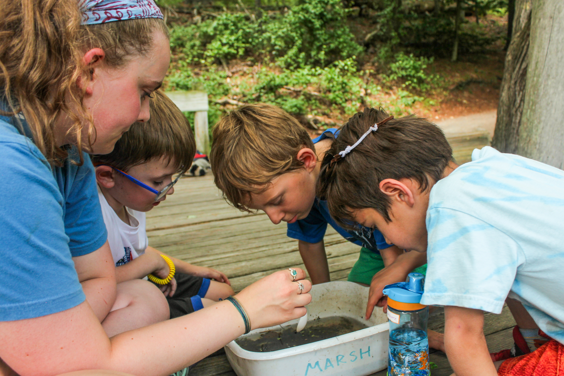 Three campers peer down into a white tub filled with water and mud that says "Marsh" on the side while a counselor gently pokes the sample with a plastic spoon