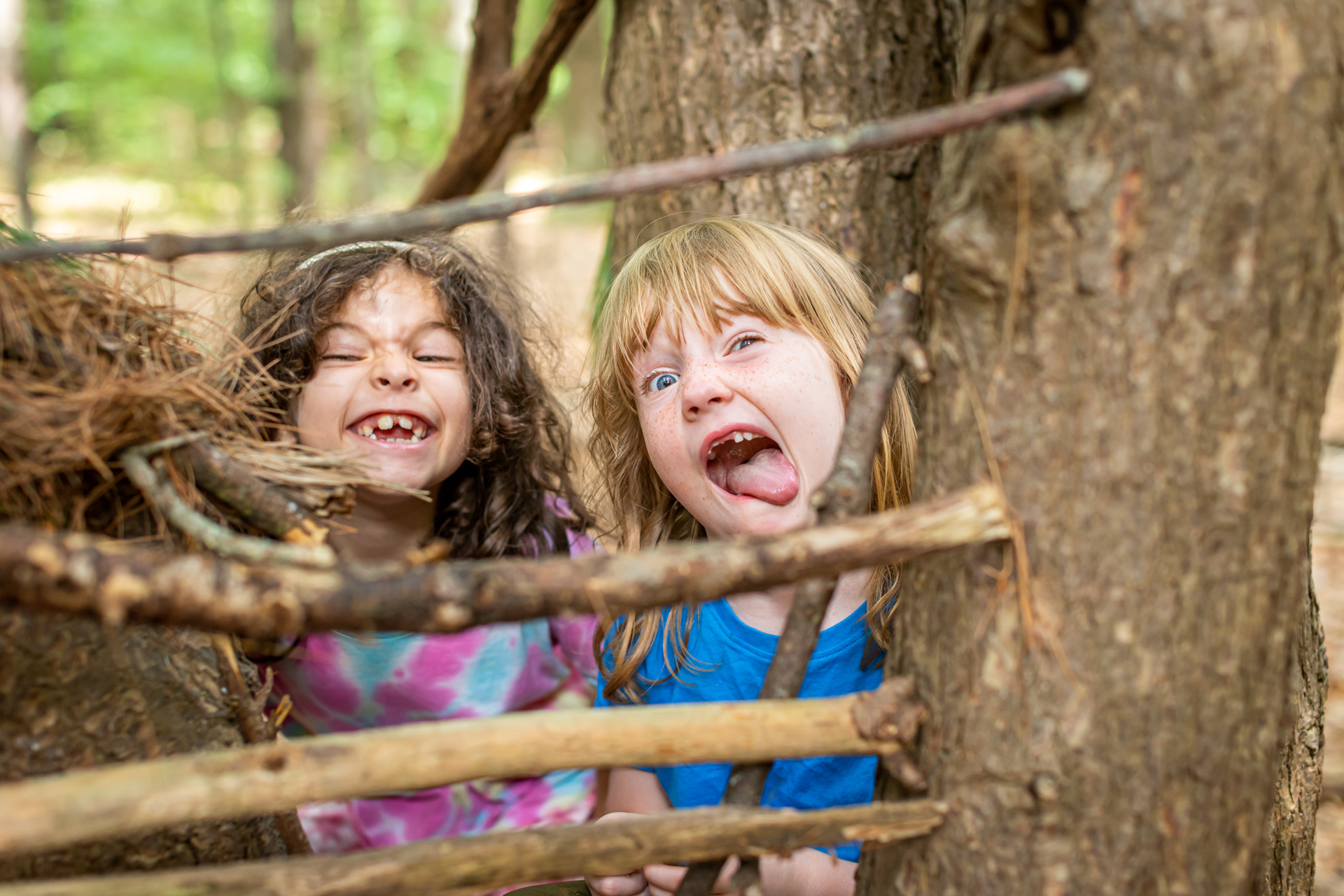 Two campers at Arcadia Nature Camp make silly faces for the camera from inside a shelter they've made using branches and twigs wedged between trees