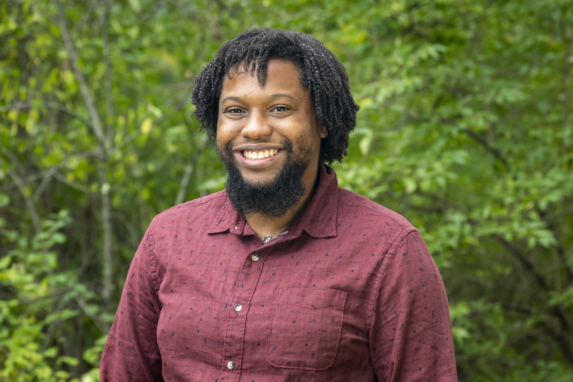 Jovan is wearing a maroon shirt and smiling with trees in the background.