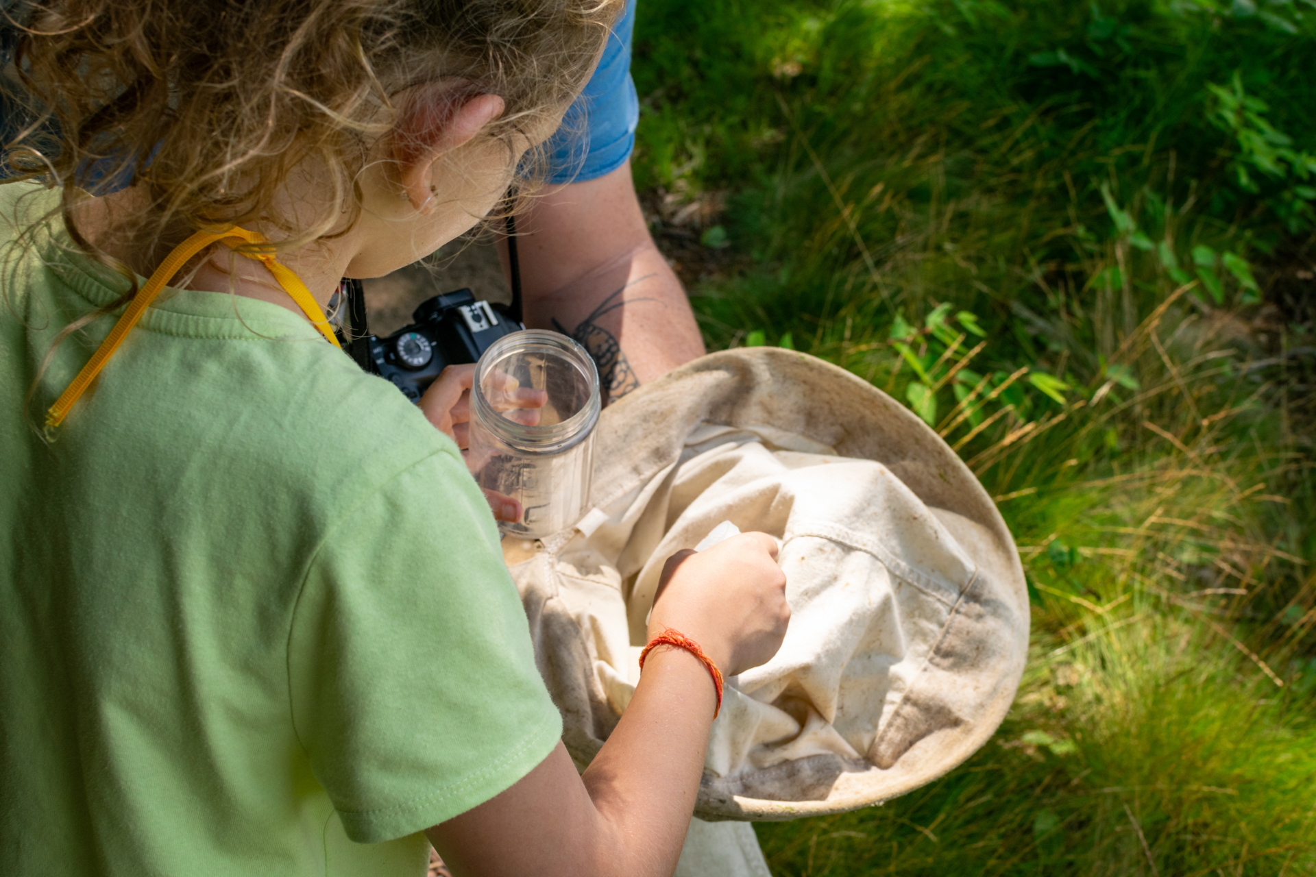 A Broadmoor camper wearing a green t-shirt reaches into a canvas bug net held by a counselor while holding a clear specimen jar in her other hand