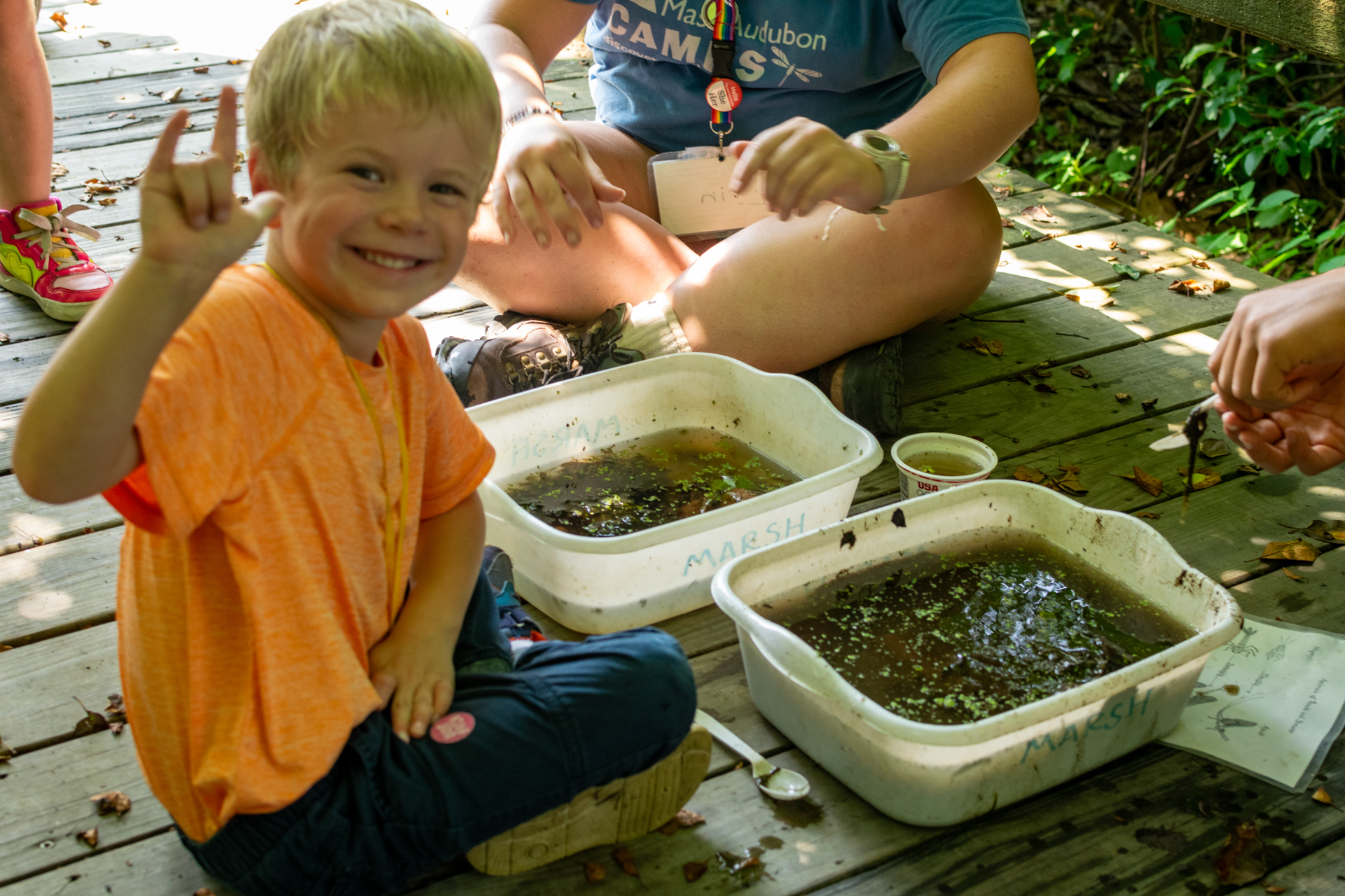 A smiling Broadmoor camper wearing an orange t-shirt sits cross-legged on a wooden boardwalk in front of two white plastic tubs containing water, mud, and aquatic plants. He is turned toward the camera and is making an "I love you" sign with his hand.