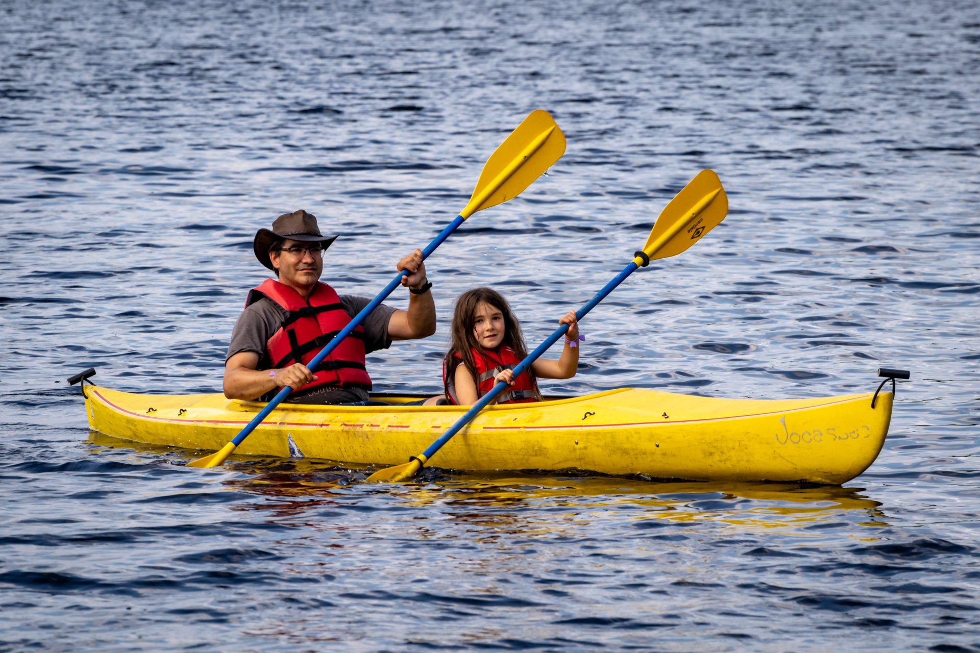 A father and daughter wearing red lifejackets are paddling a yellow, two-person kayak together.