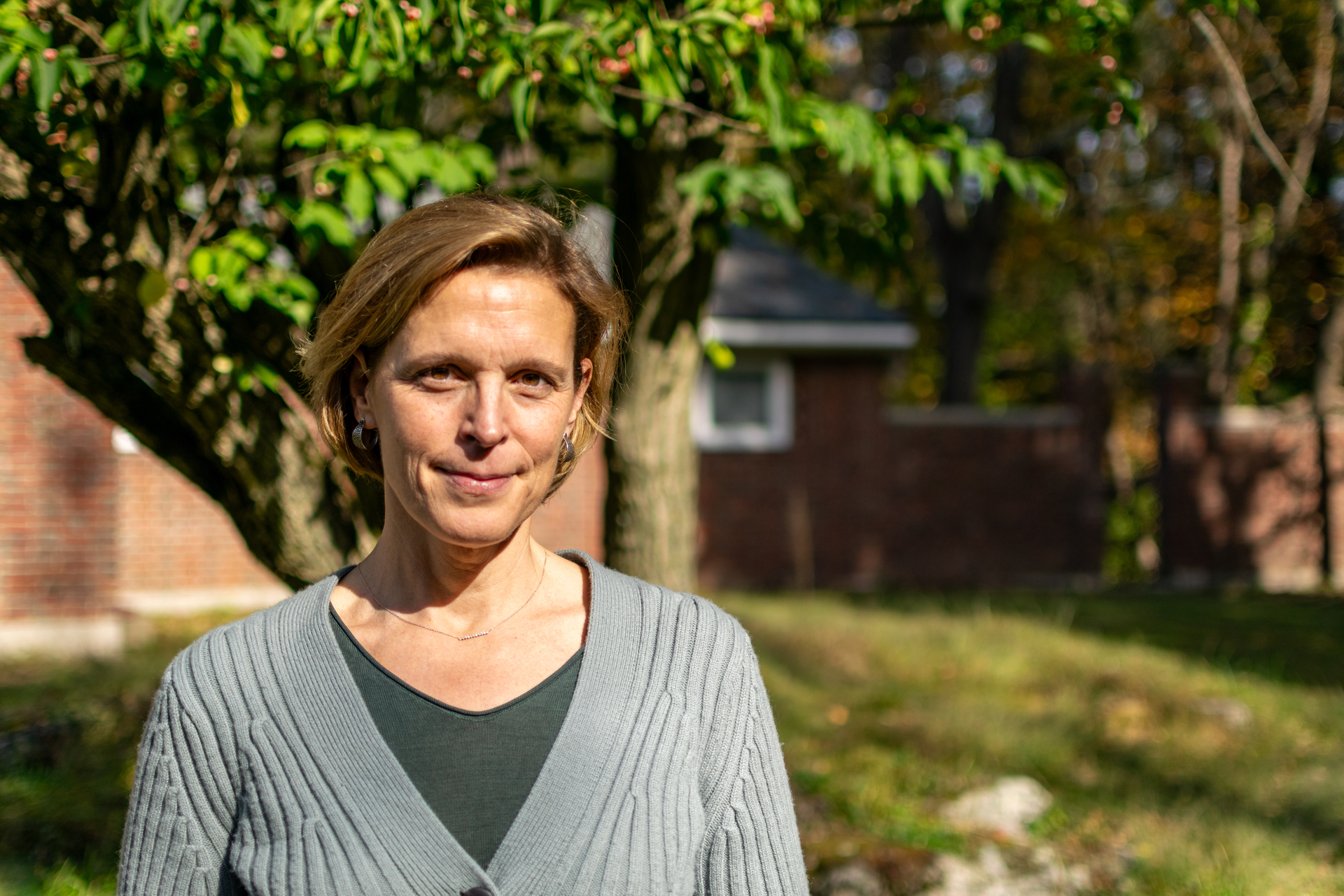 Woman with short hair smiling at the camera. Green grass and leaves are in the background.