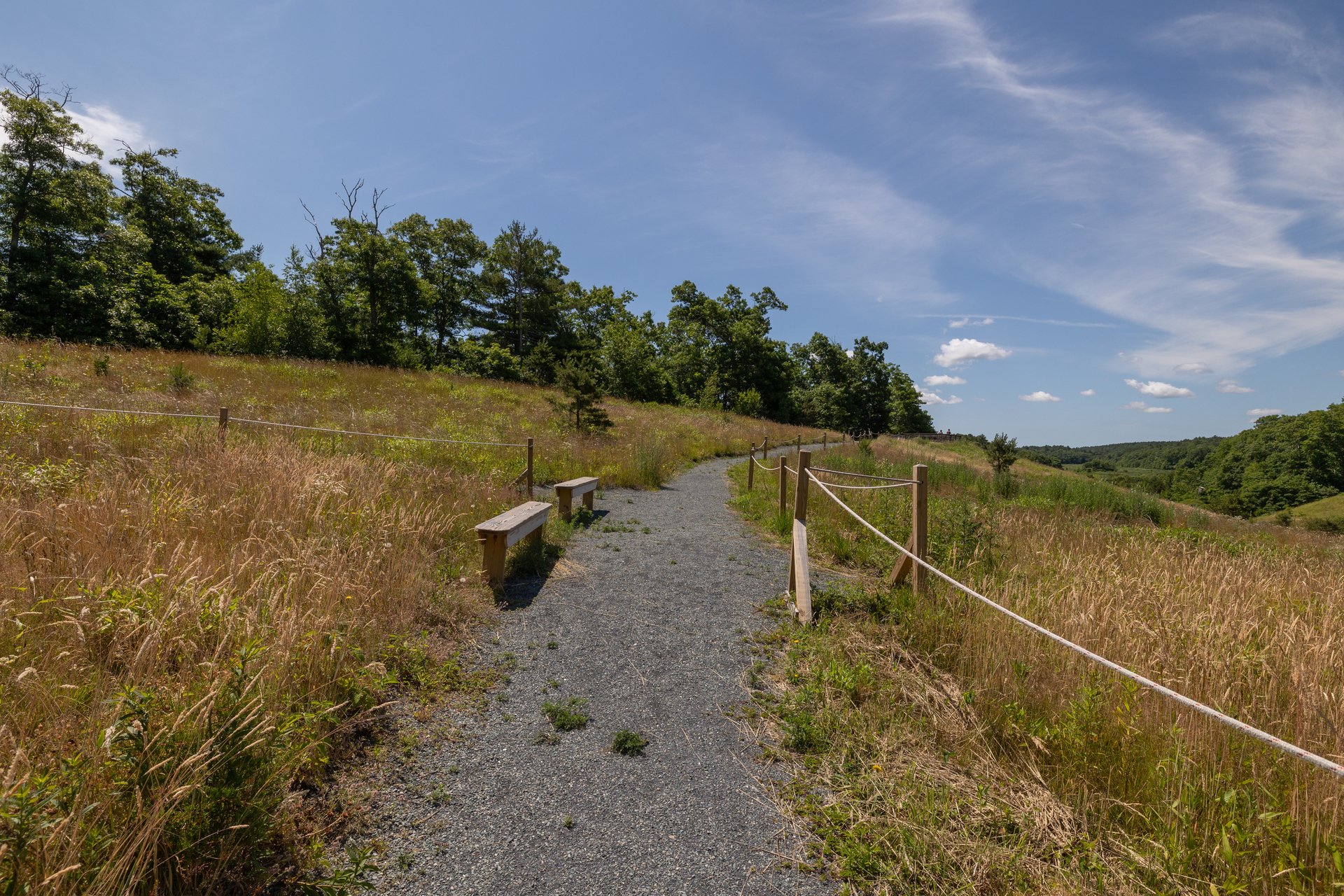 A gravel path on a hillside. On the right side of a path is a guiding rope. Two wooden benches are placed on the left.