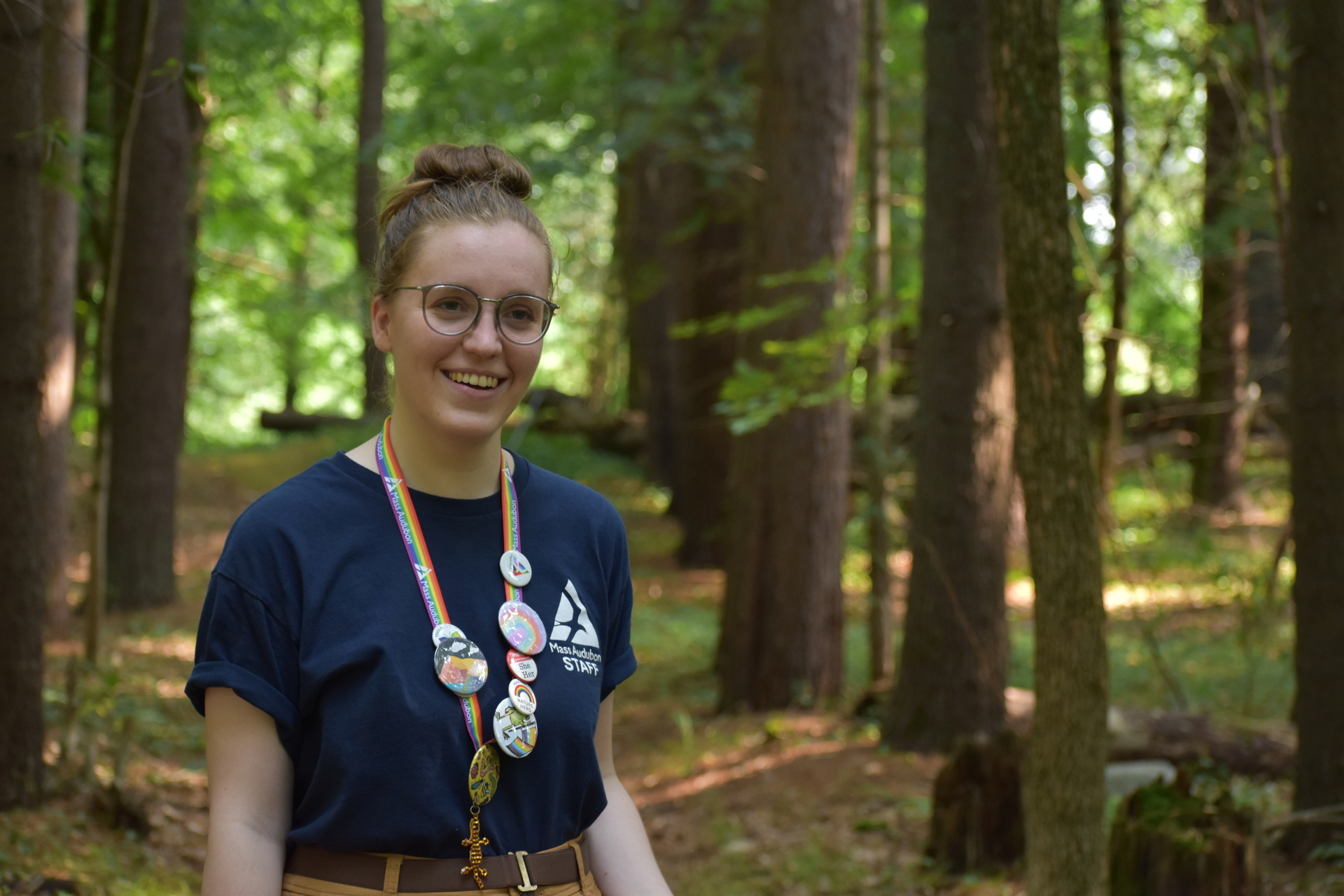 A girl with her hair in a bun standing in the woods. Around her neck is a rainbow lanyard covered in buttons.