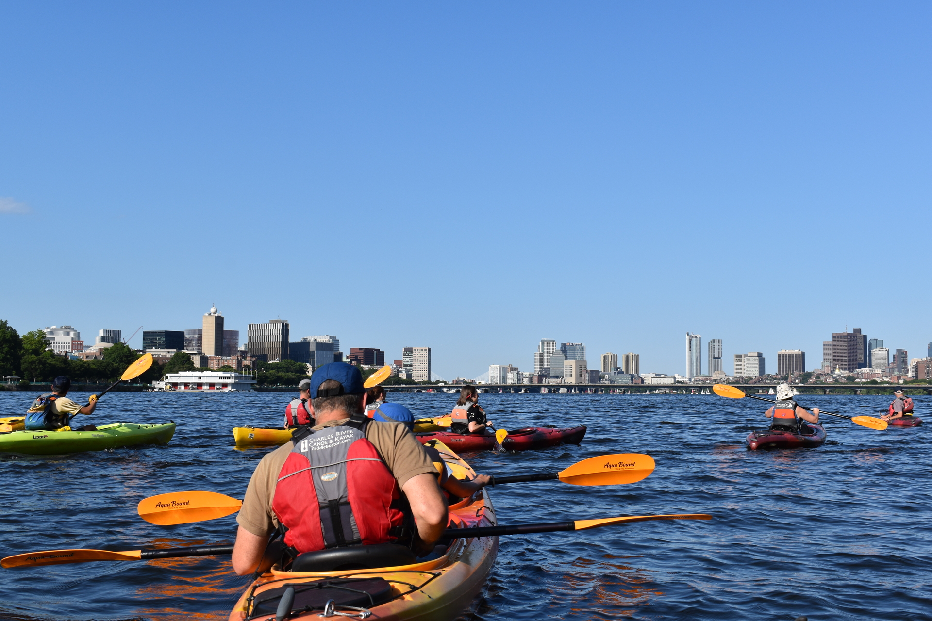 A group of kayakers in green or orange kayaks on a river. In the distance is a bridge and the Boston skyline.