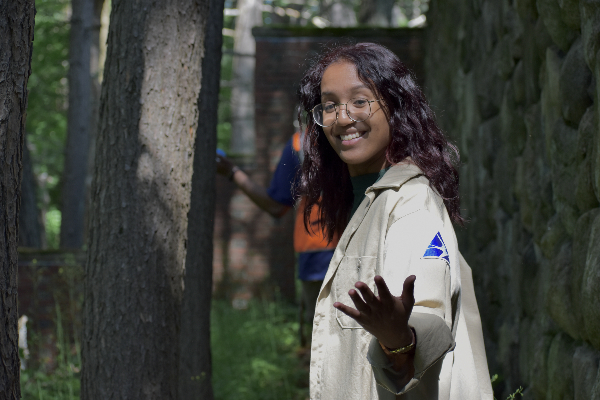 A girl with glasses wearing a tan jacket stands with her arm outstretched towards the camera. She is in a forest.