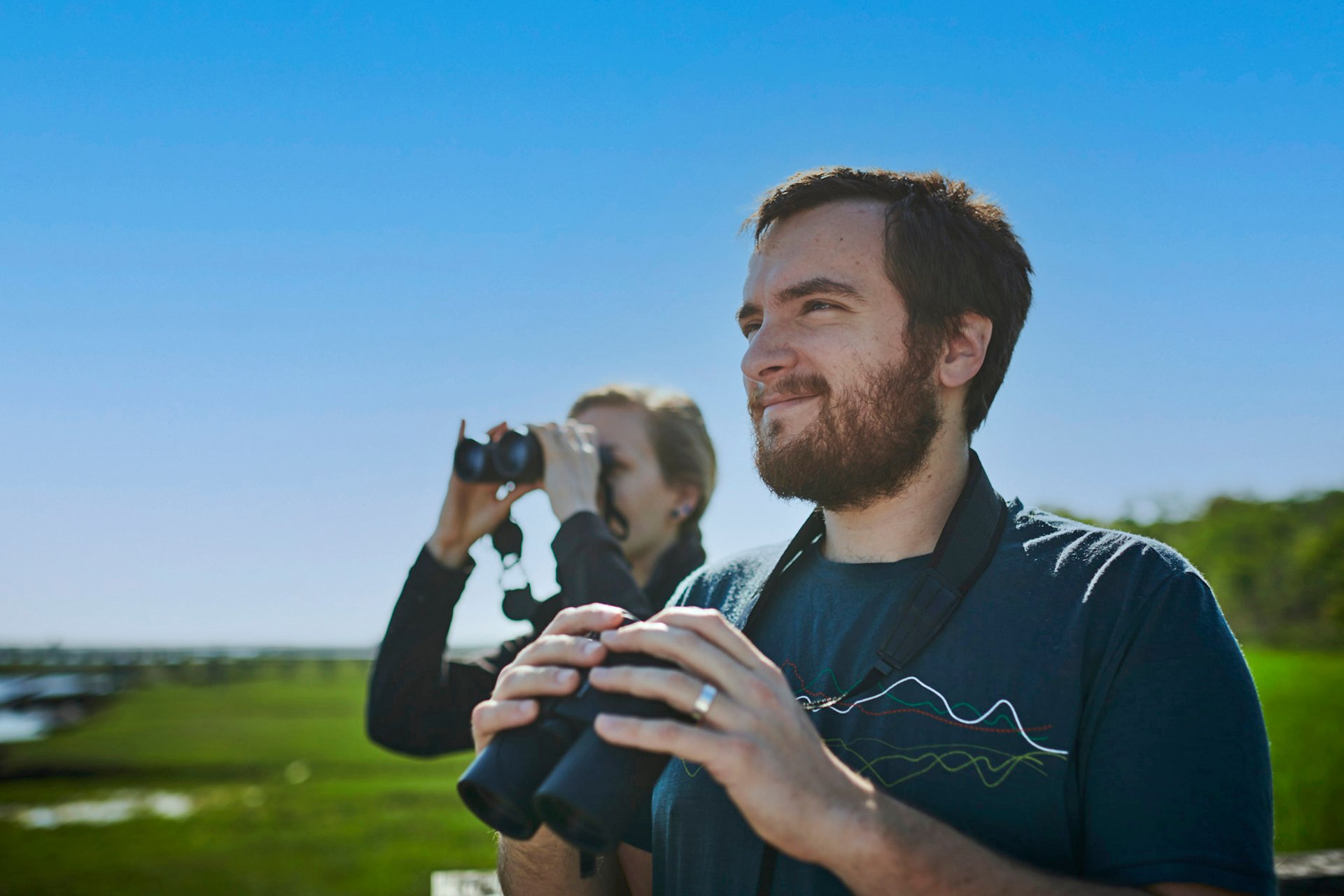 Man and Woman birding, both with binoculars, marsh behind them.