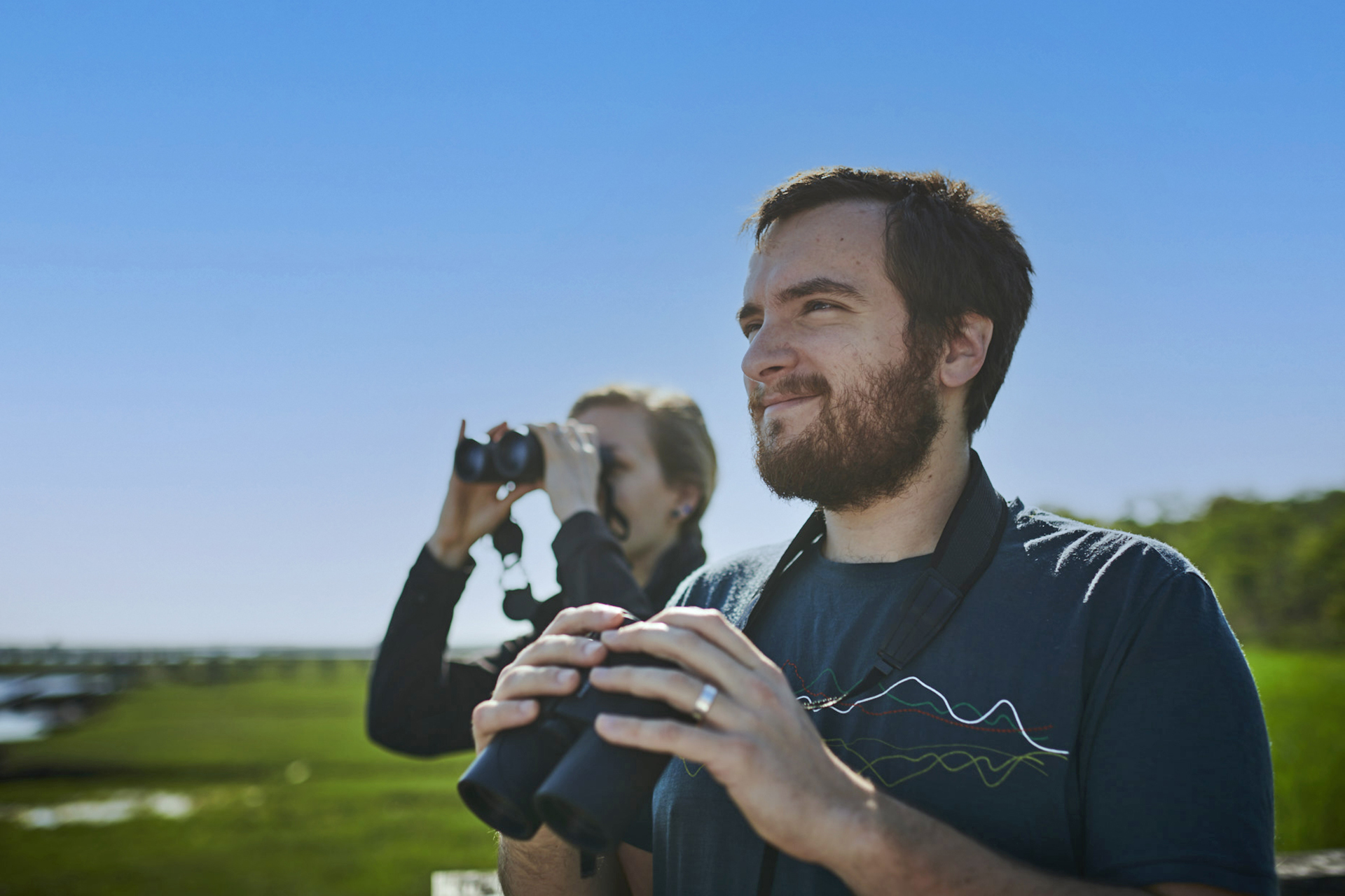 Man and Woman birding, both with binoculars, marsh behind them.