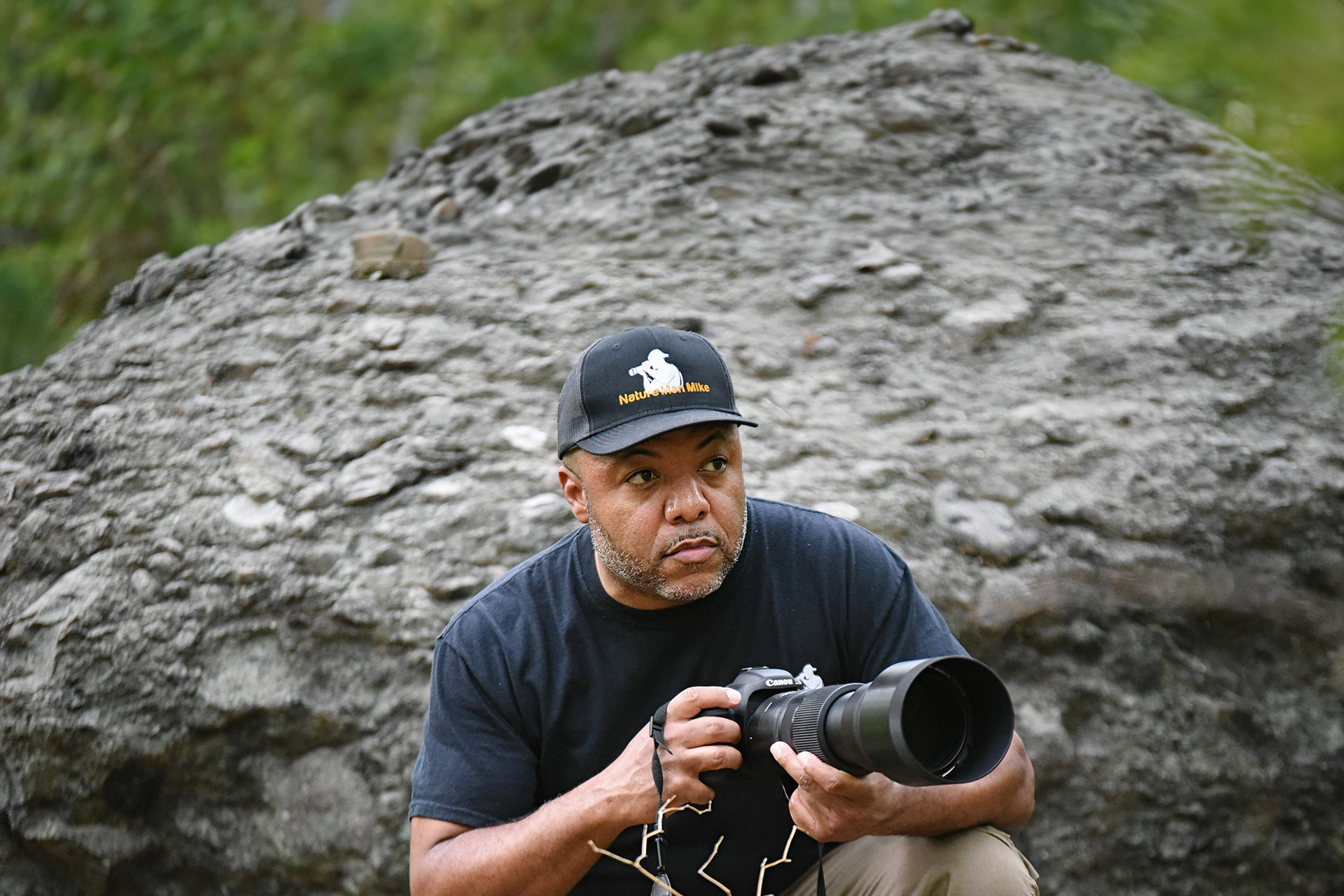 A man crouched in front of a rock with a camera.