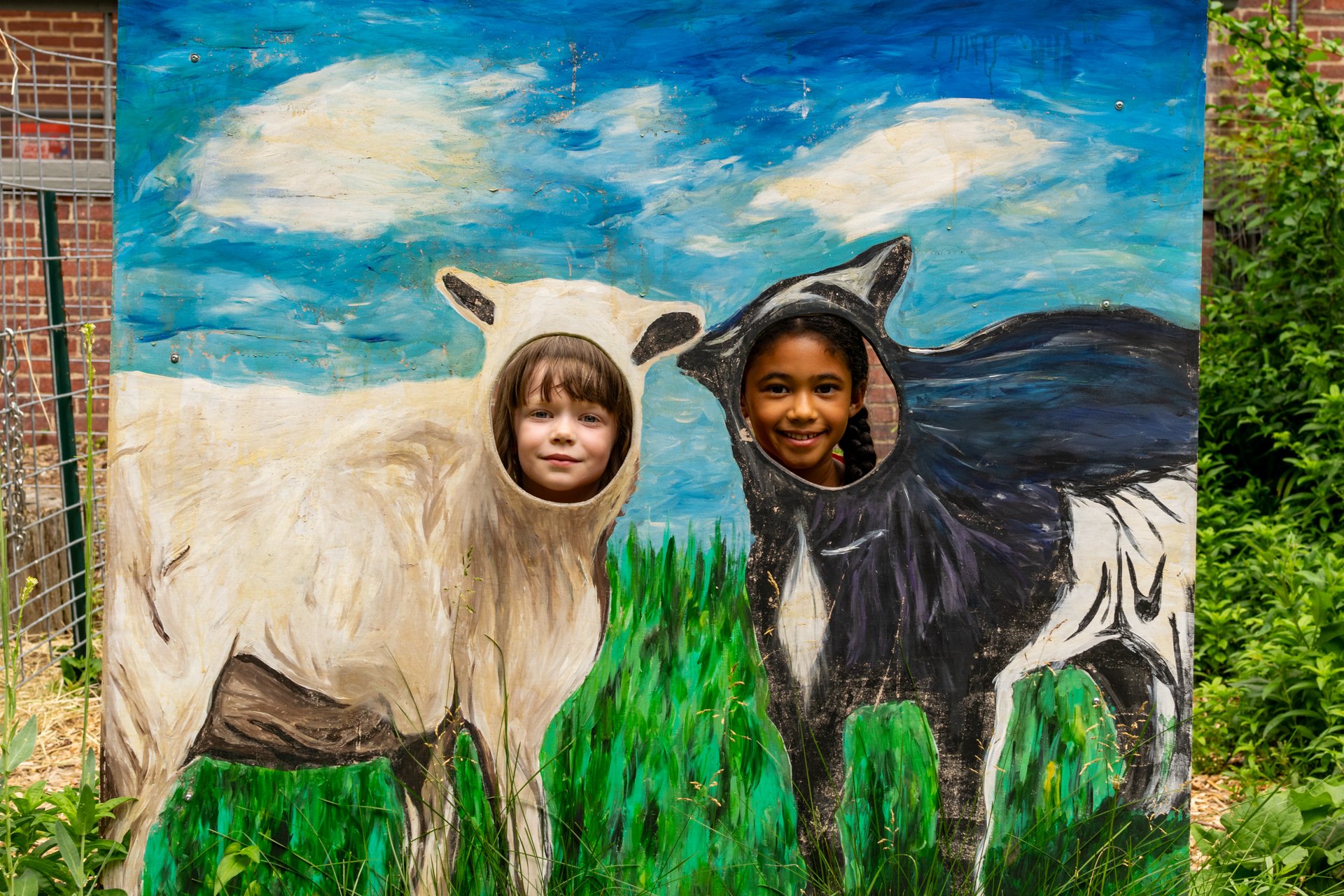 Two habitat campers pose for a photo behind a large painting of two goats with cutout circles so the kids can poke their faces through