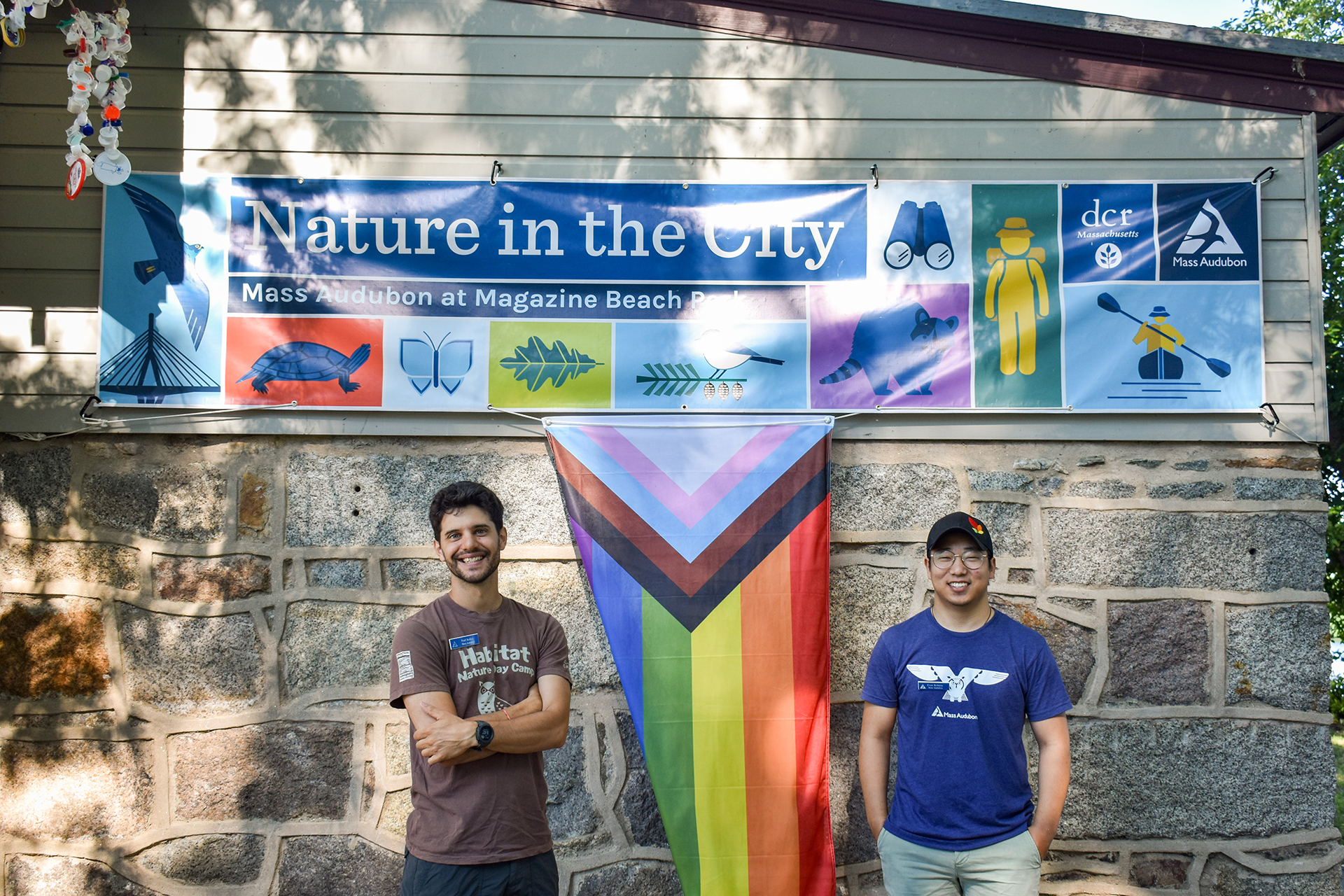 Two young men standing under a banner, with the Progress Pride flag hanging from it, framed between them