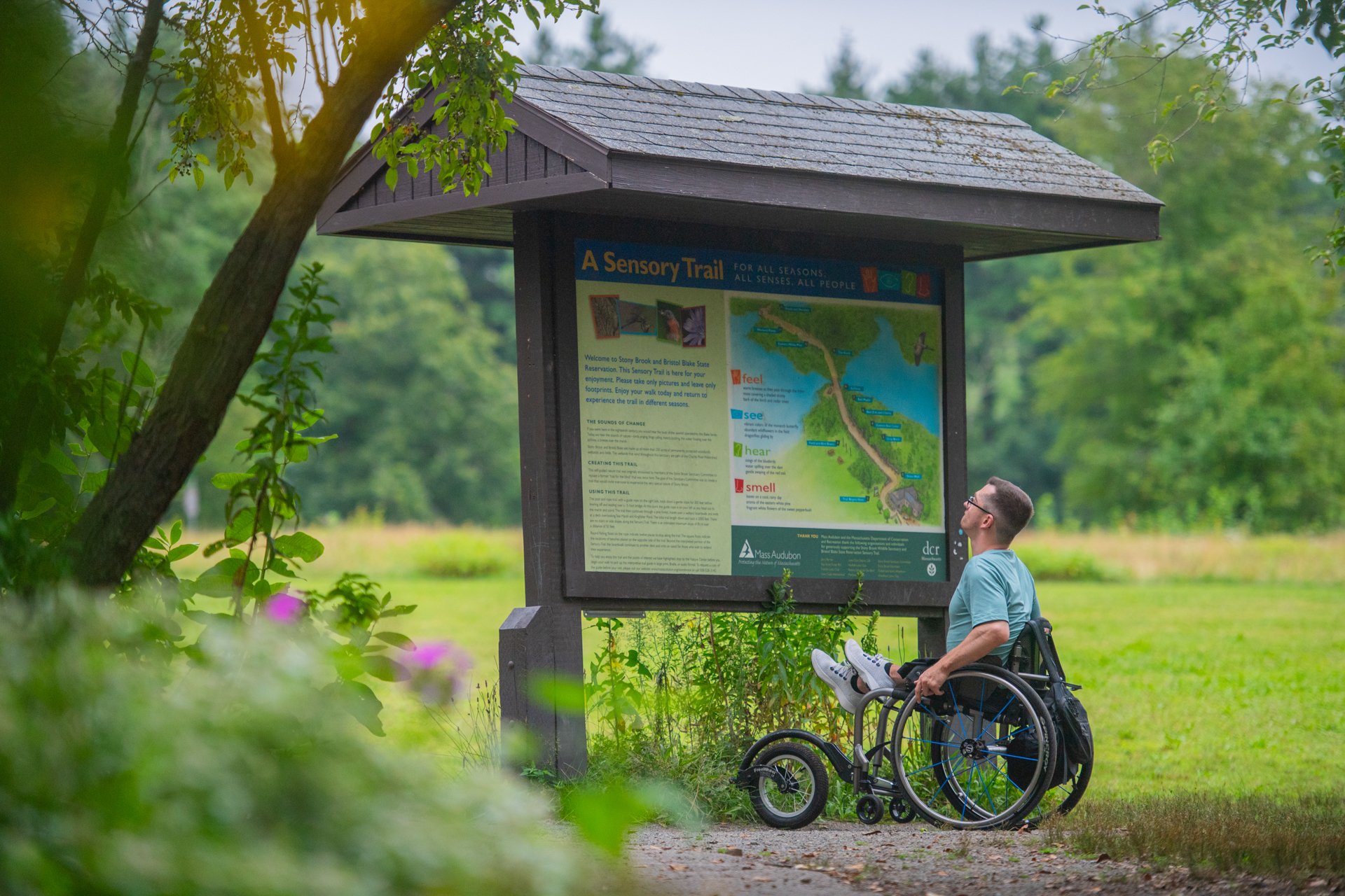A person using a wheelchair reading an interpretive sign with a map at a trail kiosk on Stony Brook's accessible All Persons Trail