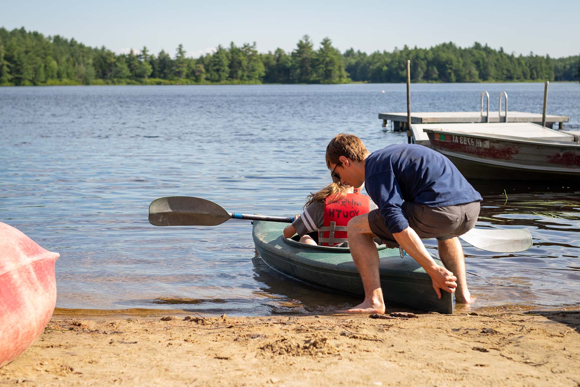 Person bent down pushing a kayak into the lake with a person in it.