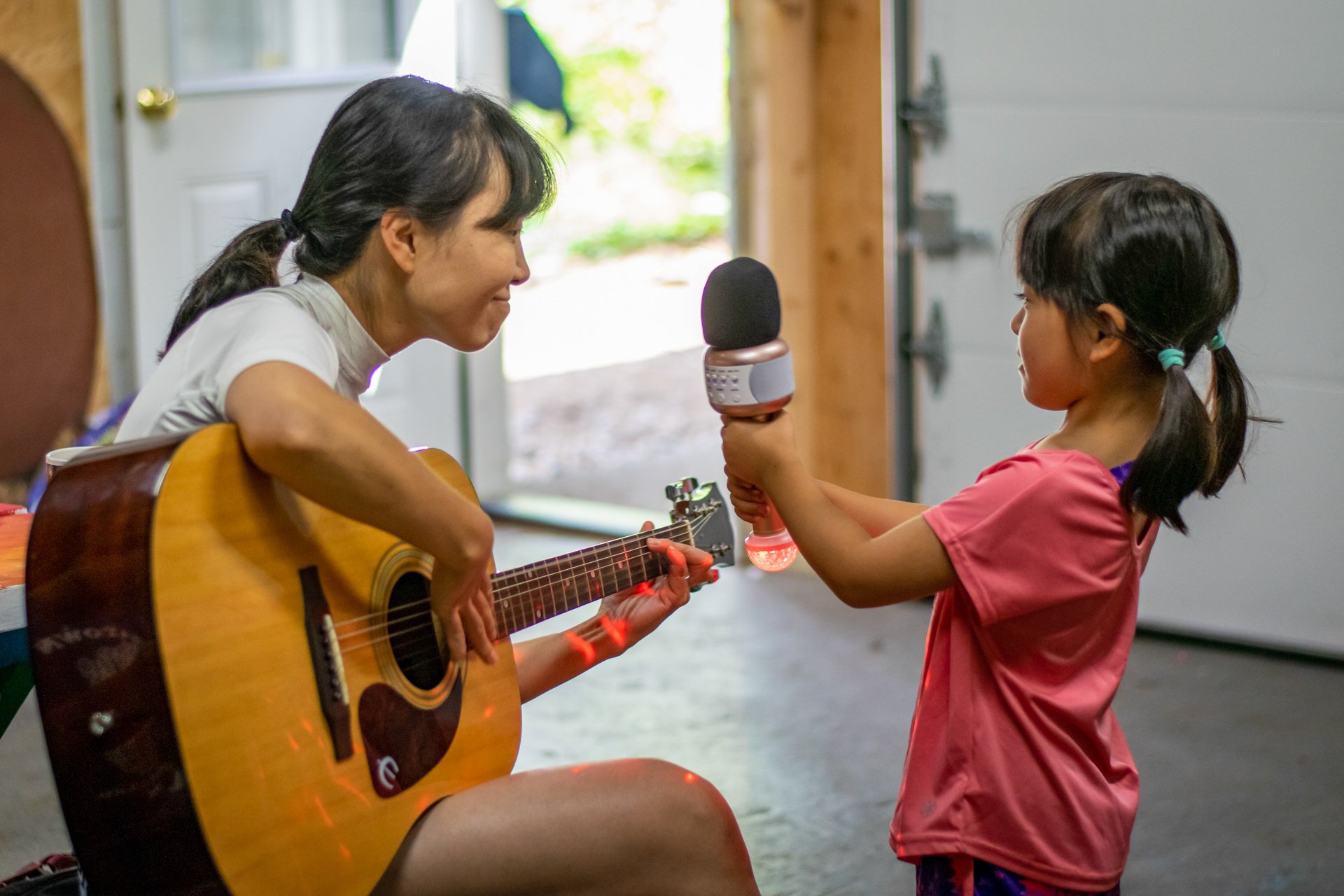 A mom plays an acoustic guitar while her daughter holds up fake microphone for her to sing into